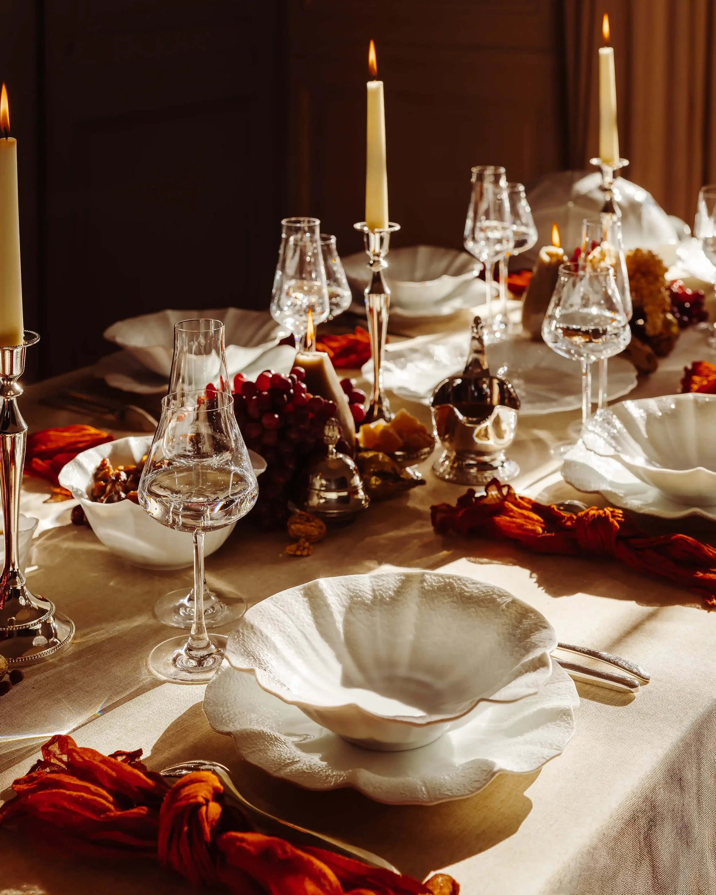 Elegant dining table set with white plates, crystal wine glasses, silver candle holders with lit candles, and decorative flowers, in a warmly lit room.