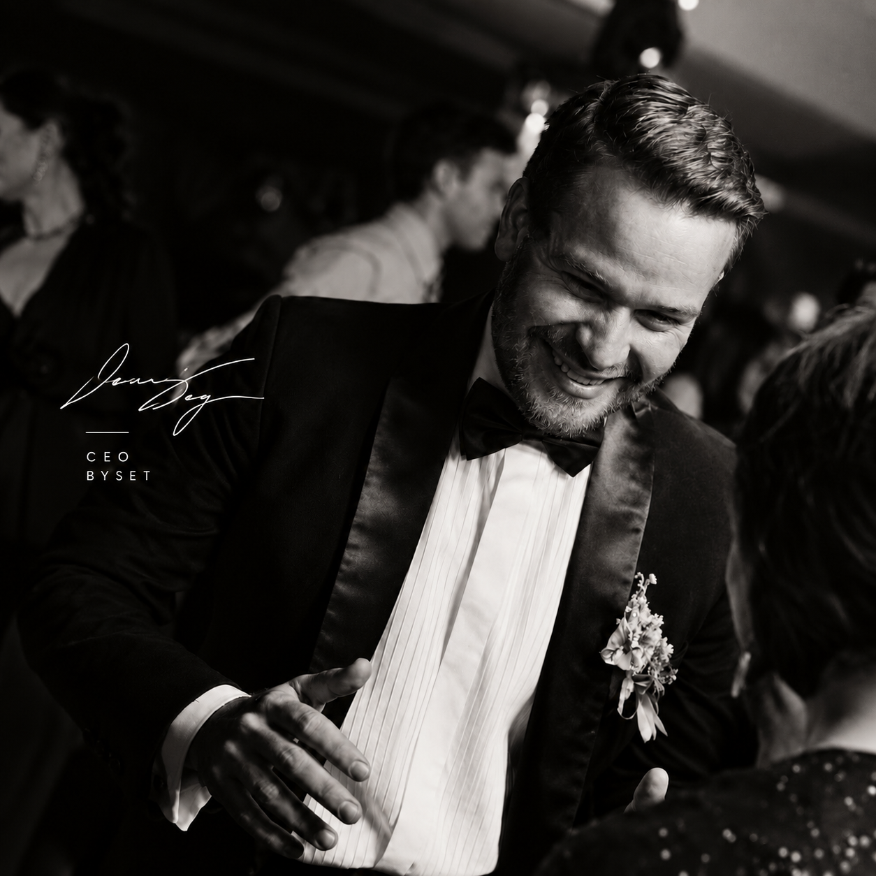 A black and white photo of a man in a tuxedo smiling and talking to a woman at a formal event.