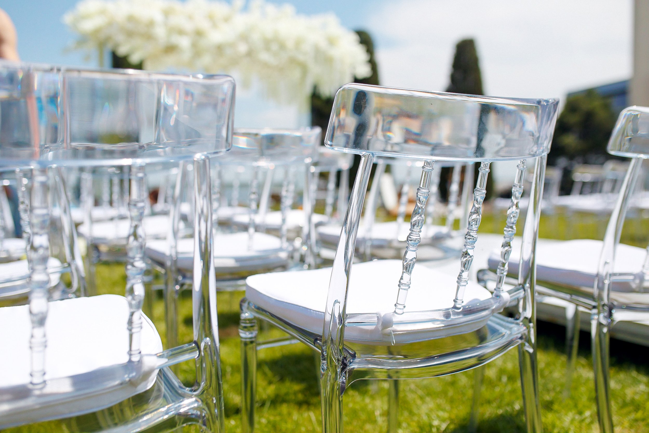 Clear acrylic chairs arranged outdoors on green grass for an event with a blurred background of trees and white flowers.