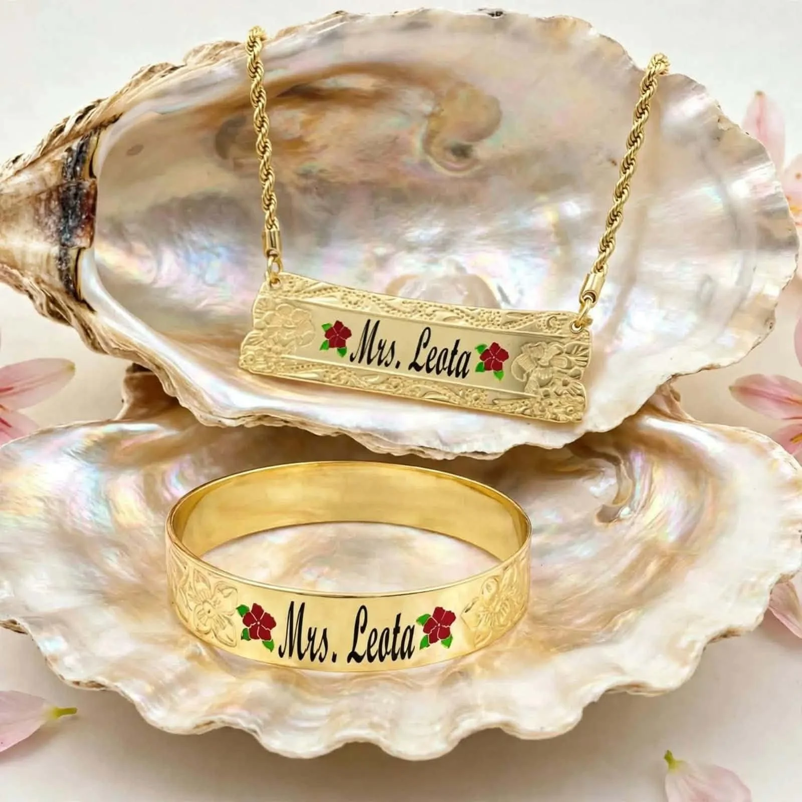 A gold bracelet with the inscription 'Mrs. Leata' and red flower decorations, resting inside a large iridescent shell with a matching necklace above it. The shell is displayed against a soft, light-colored background with pink flower petals.