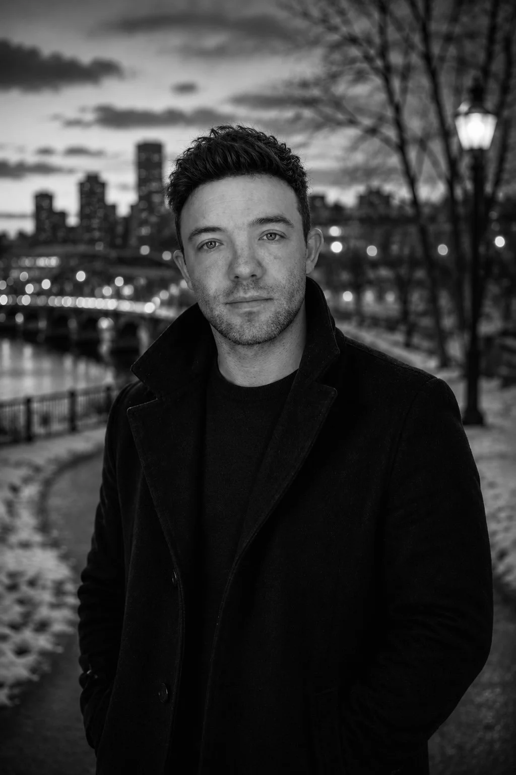 A black-and-white photo of a man with short dark hair, standing outdoors near a river at dusk with city skyline and trees in the background.