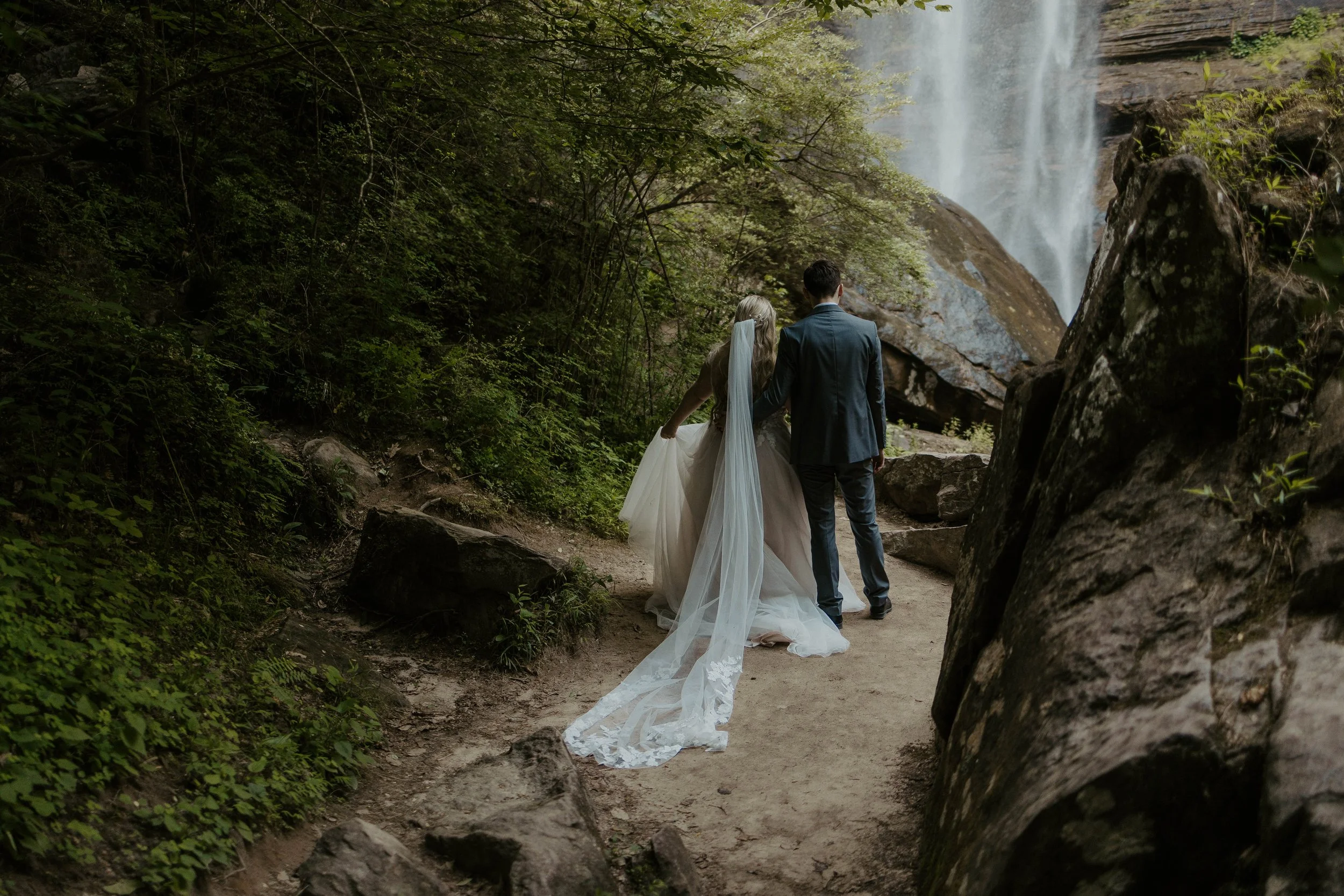A bride and groom walking on a forest trail near a waterfall, with lush greenery and rocks surrounding them.