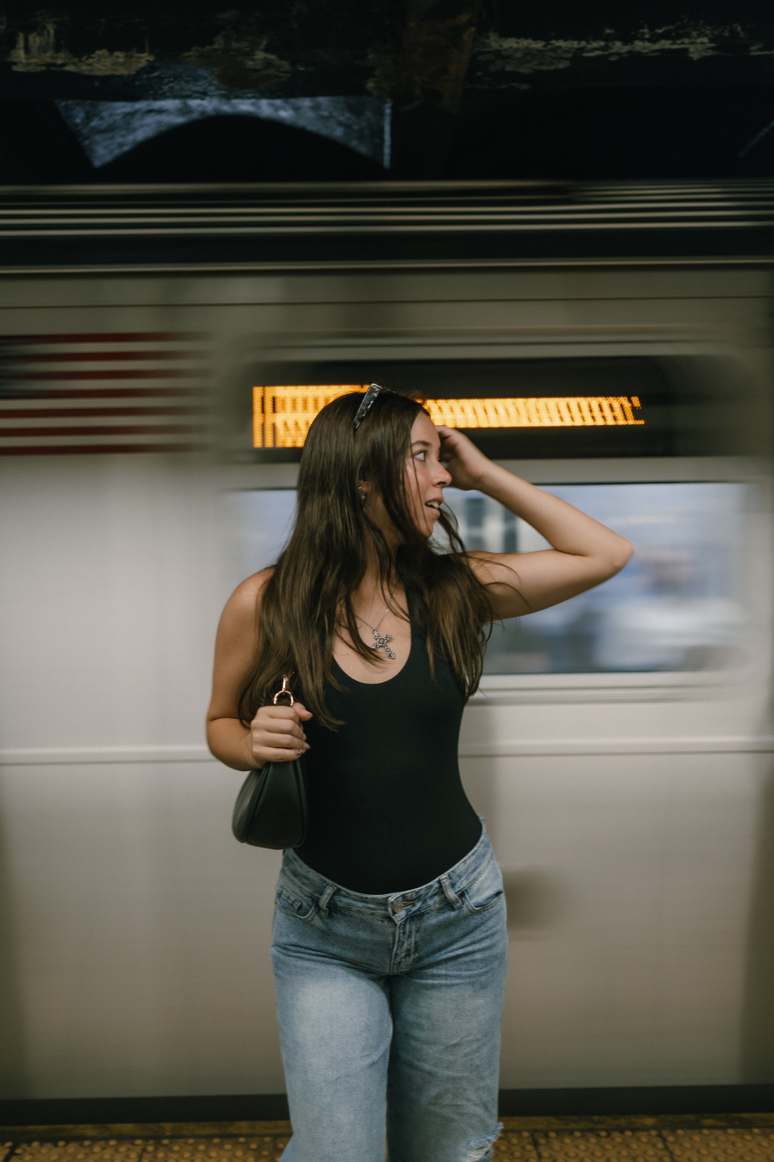 Young woman with long brown hair, wearing a black tank top, light blue jeans, black sunglasses on her head, and holding a black purse, standing in front of a moving train at a train station.