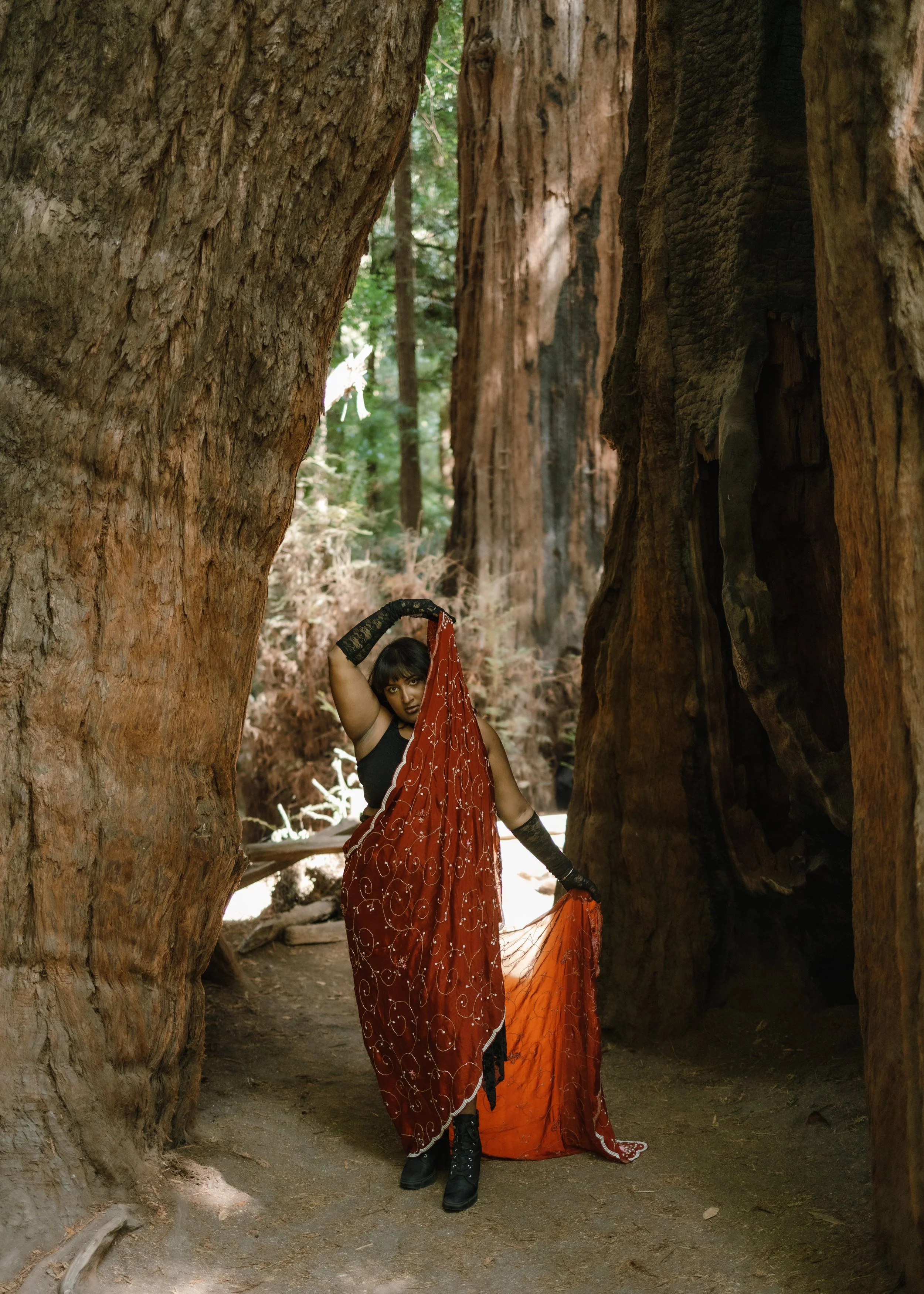 A woman wearing a black top, black boots, black fingerless gloves, and an orange and black skirt, poses among tall trees in a forest. She holds a piece of orange fabric with white patterns, with her right arm raised above her head, and her left arm d