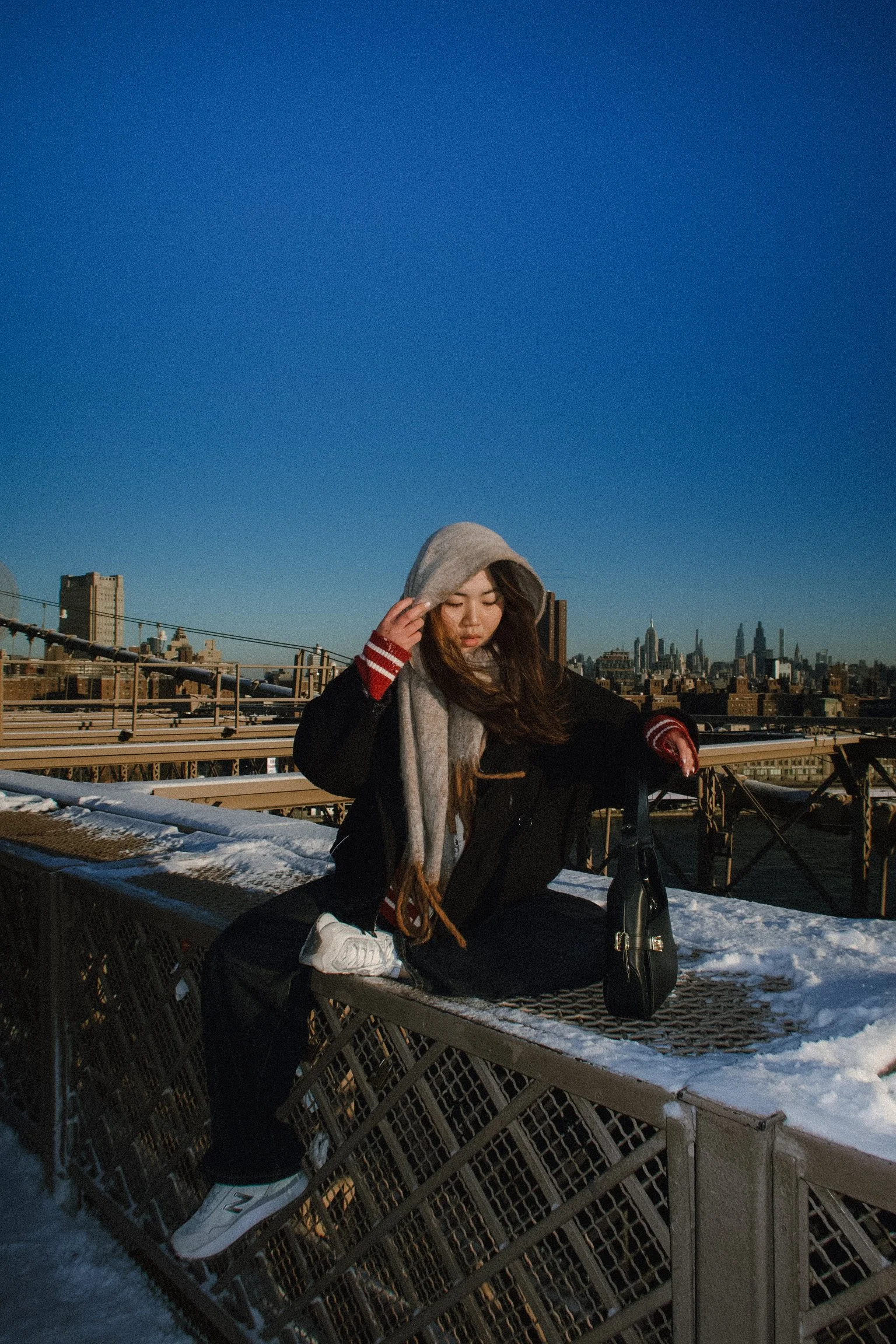 Young woman with long brown hair sitting on a metal bench on a rooftop in New York City during winter, wearing a gray hoodie and red striped sweater, with snow on the ground and city skyline in the background under a clear blue sky.