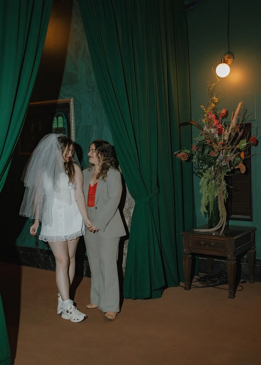 Two women holding hands and smiling in front of dark green curtains, one wearing a white dress with a veil and sneakers, the other wearing a light-colored suit with a red top, in a room with a large floral arrangement on a small table and a wall-moun