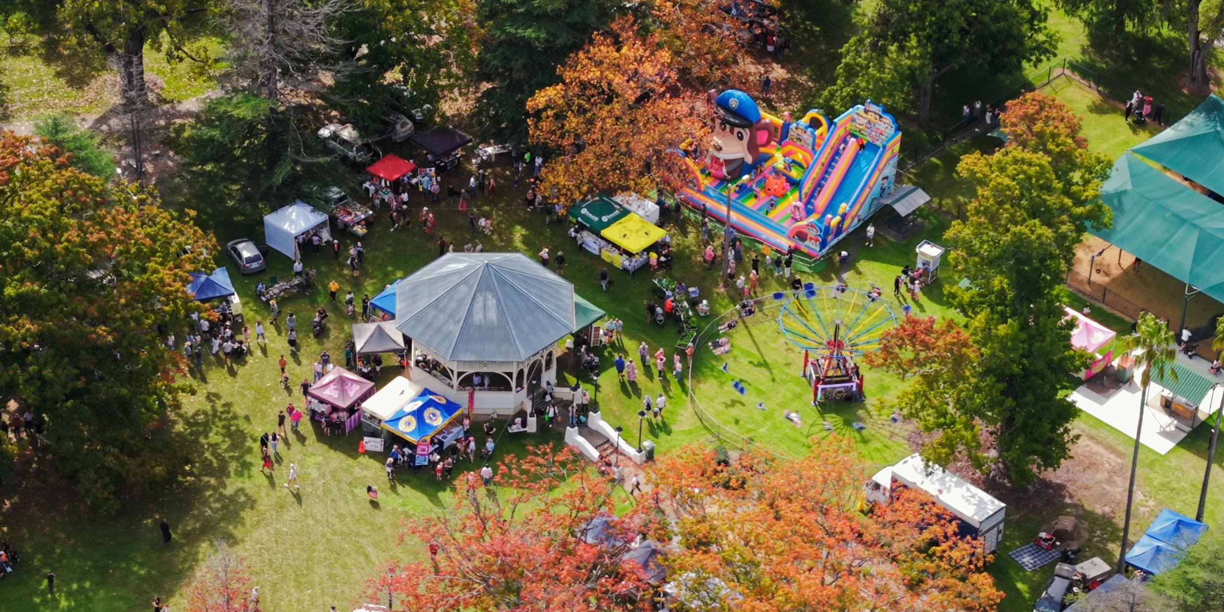 Aerial view of a crowded outdoor festival in a leafy park with vendor tents, food stalls, a gazebo, inflatable slides, and carnival rides.