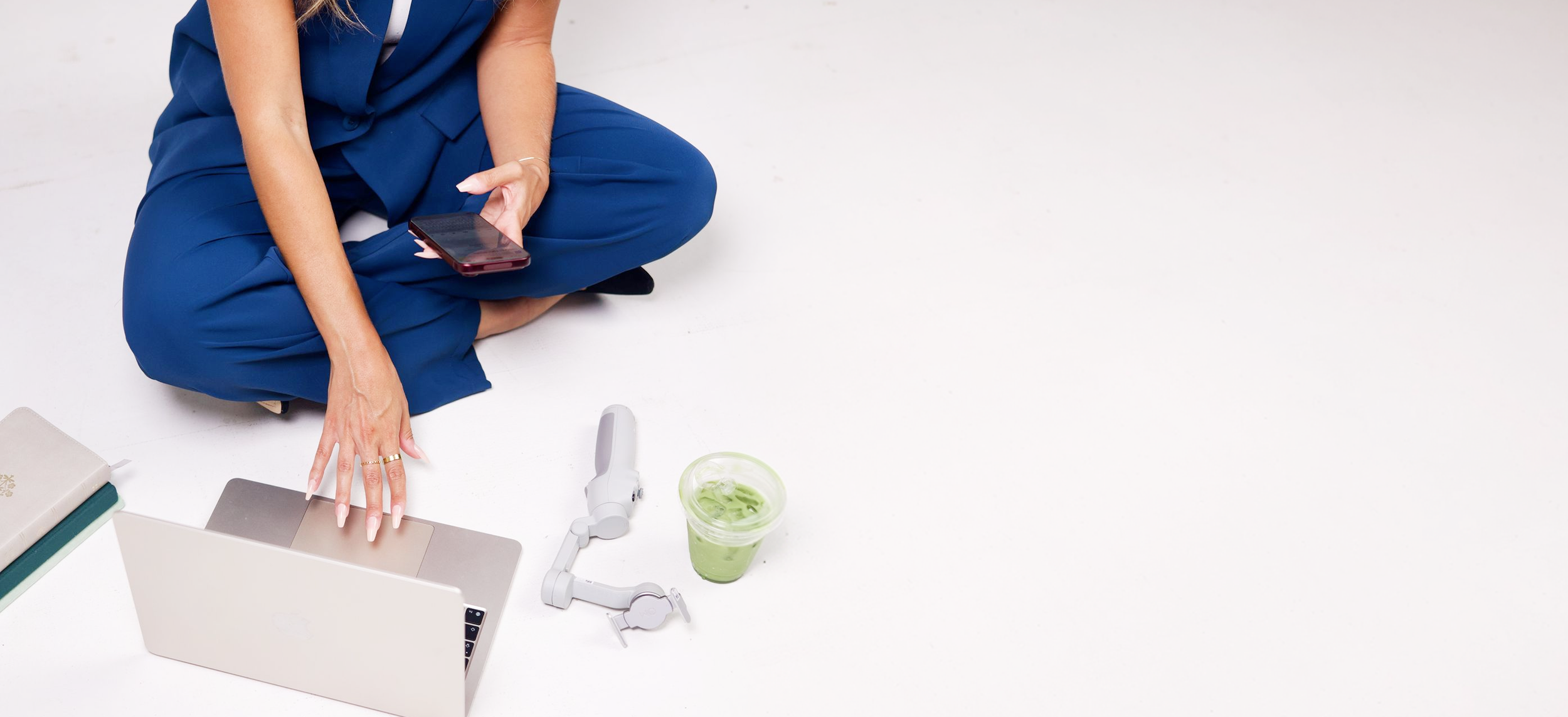 Founder Hannah Kavy sitting on the floor working on a laptop with with a phone, gimbal, notebooks and iced matcha