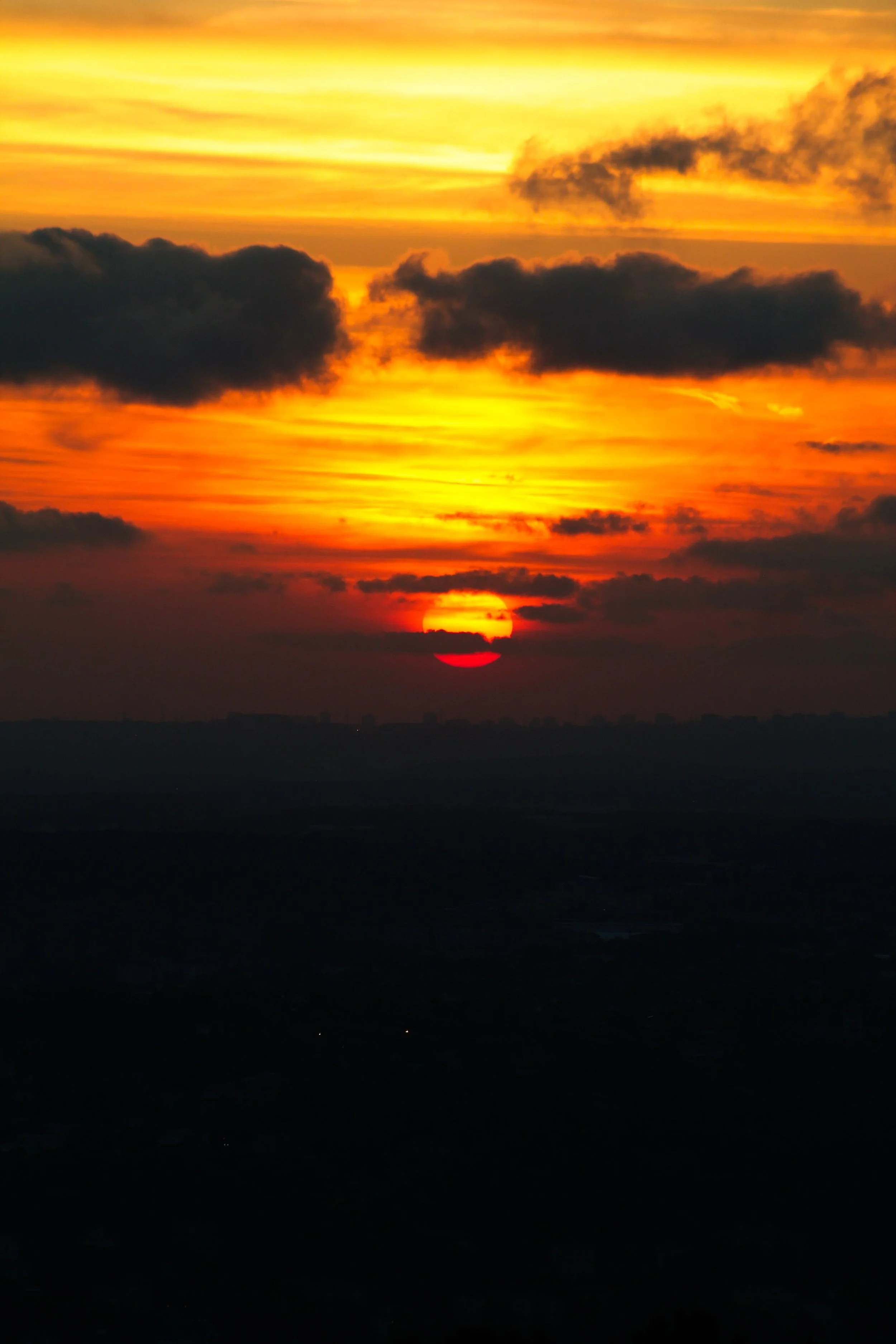 A sunset with the sun partially below the horizon, and clouds illuminated in shades of orange, yellow, and dark gray, seen over a distant landscape.
