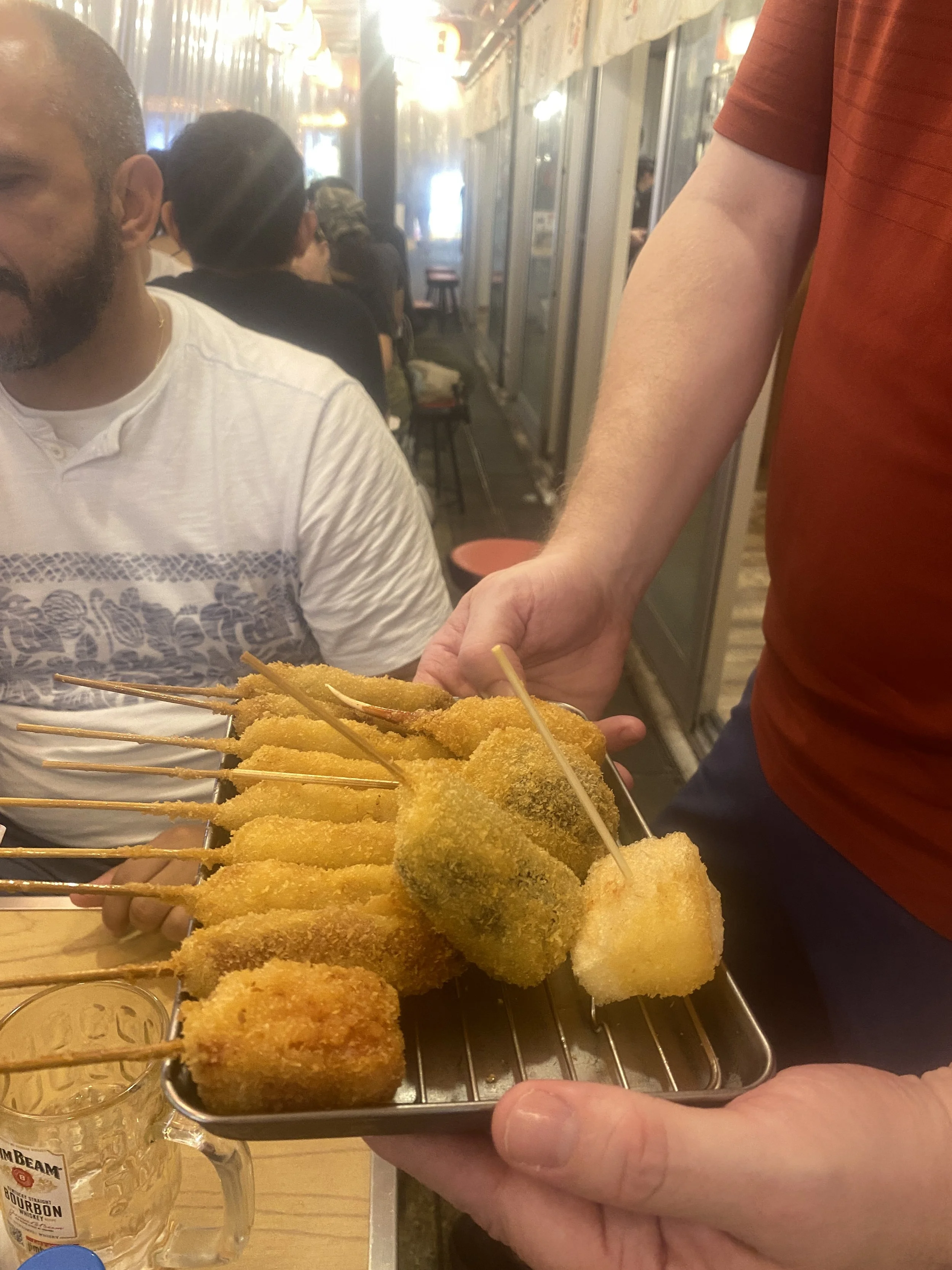 The tour guide is holding a tray of Kushikatsu (skewered fried food) in a restaurant in Shinsekai. The background shows other customers seated at tables, with warm lighting and a casual dining atmosphere.