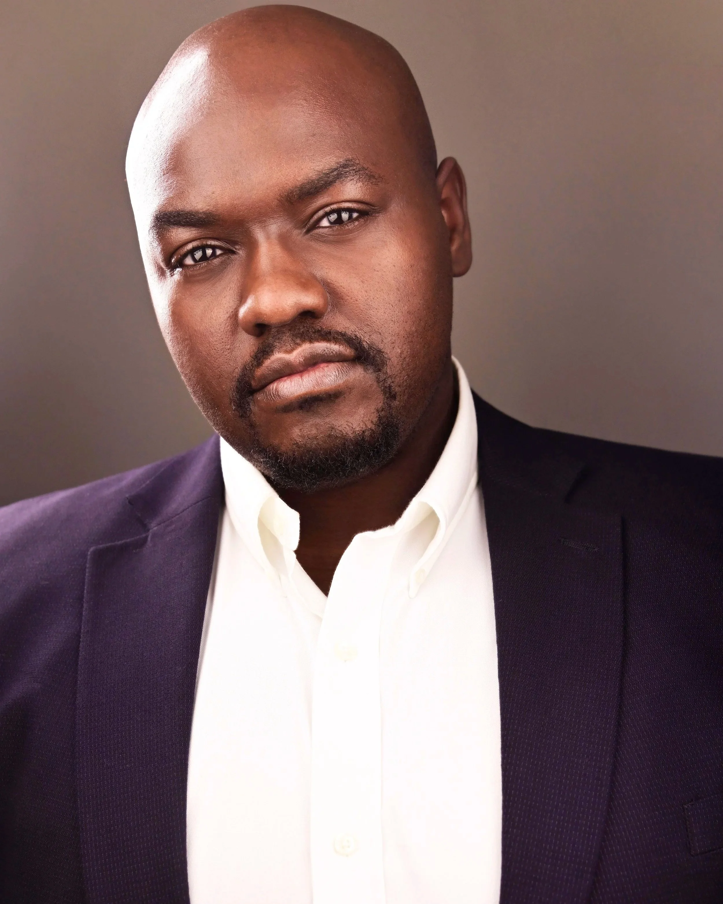 Close-up portrait of a bald Black man with a beard wearing a dark suit and white dress shirt.