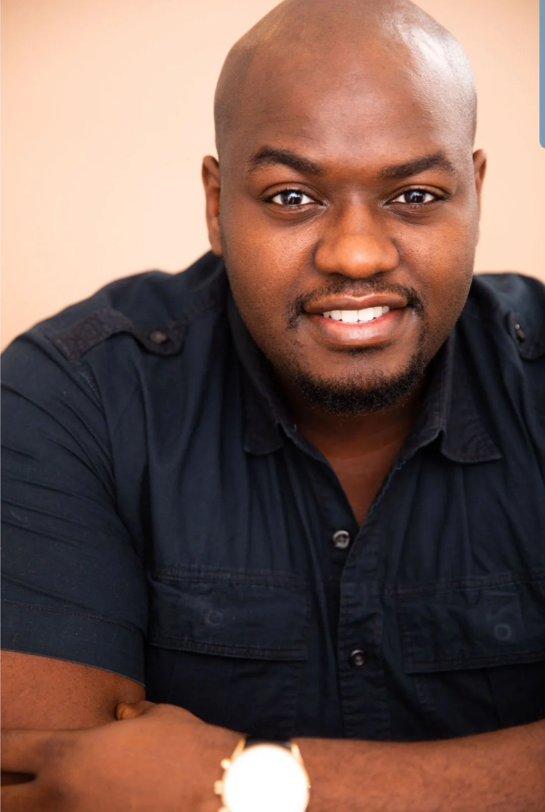 Smiling bald Black man with a short beard wearing a dark shirt, posed against a peach background.