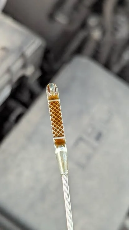 Close-up of an automotive dipstick with brown engine oil, held over a blurred engine compartment background.