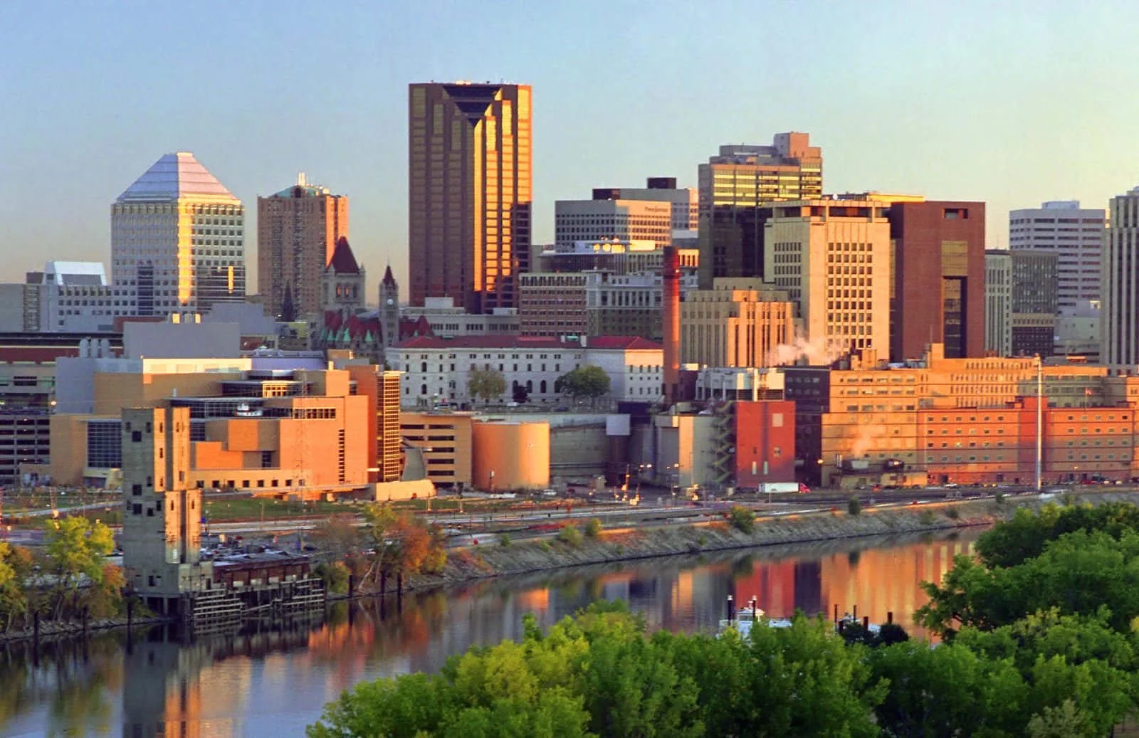 Dusk view of downtown Indianapolis skyline with a river in the foreground, modern high-rise buildings, and historic structures, reflecting on the water.