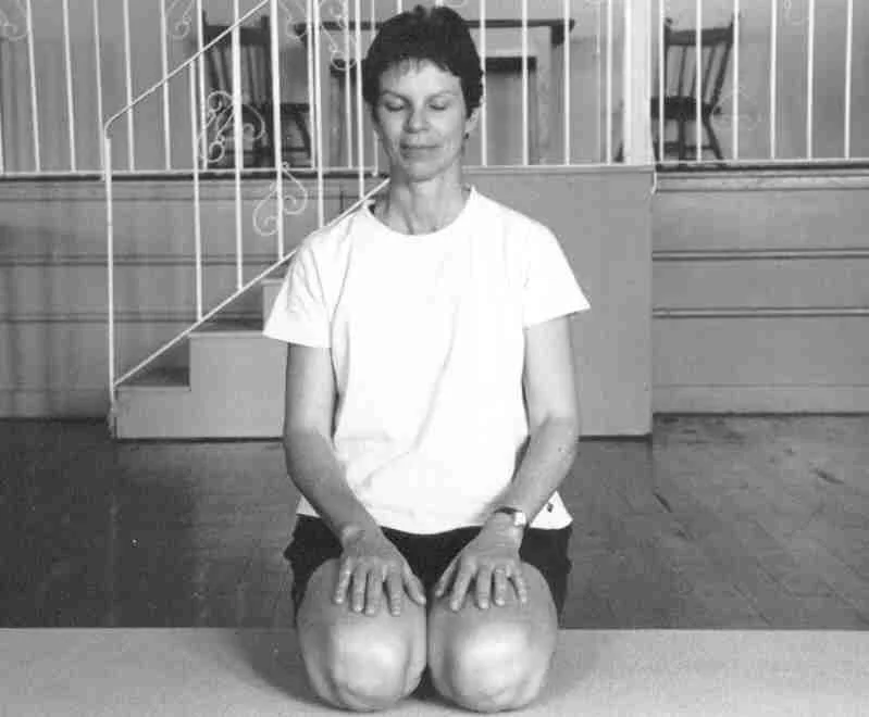 A woman kneeling on the floor with her hands resting on her thighs, in front of a staircase with a metal railing, in a room with wooden floors and bookshelves in the background.