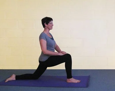 A woman performing a kneeling lunge yoga pose on a purple mat against a yellow wall.