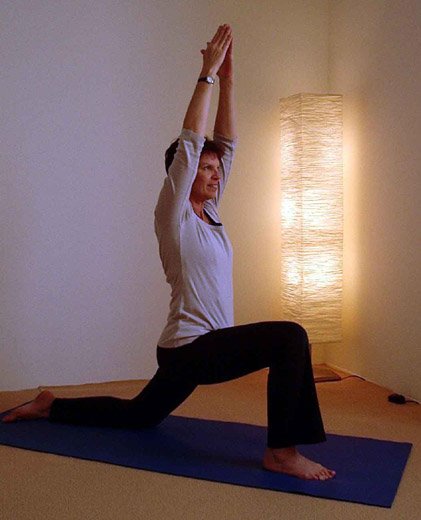 Woman practicing yoga in a lunge position with hands raised above her head, indoors near a lit floor lamp.