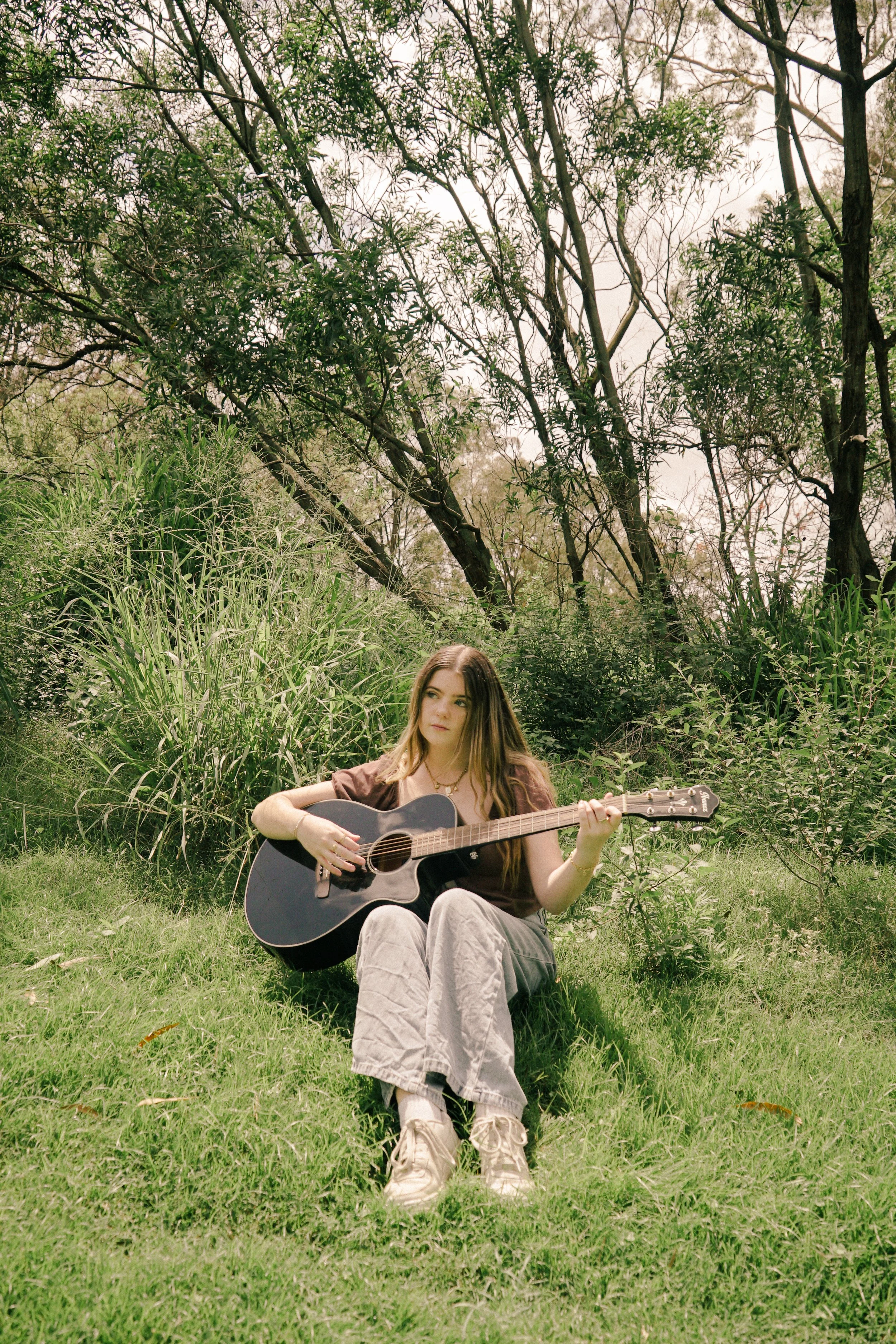 A young woman sitting on the grass outdoors playing an acoustic guitar. She has long hair, and is dressed casually in a brown top, beige pants, and sneakers. The background features lush greenery and trees.