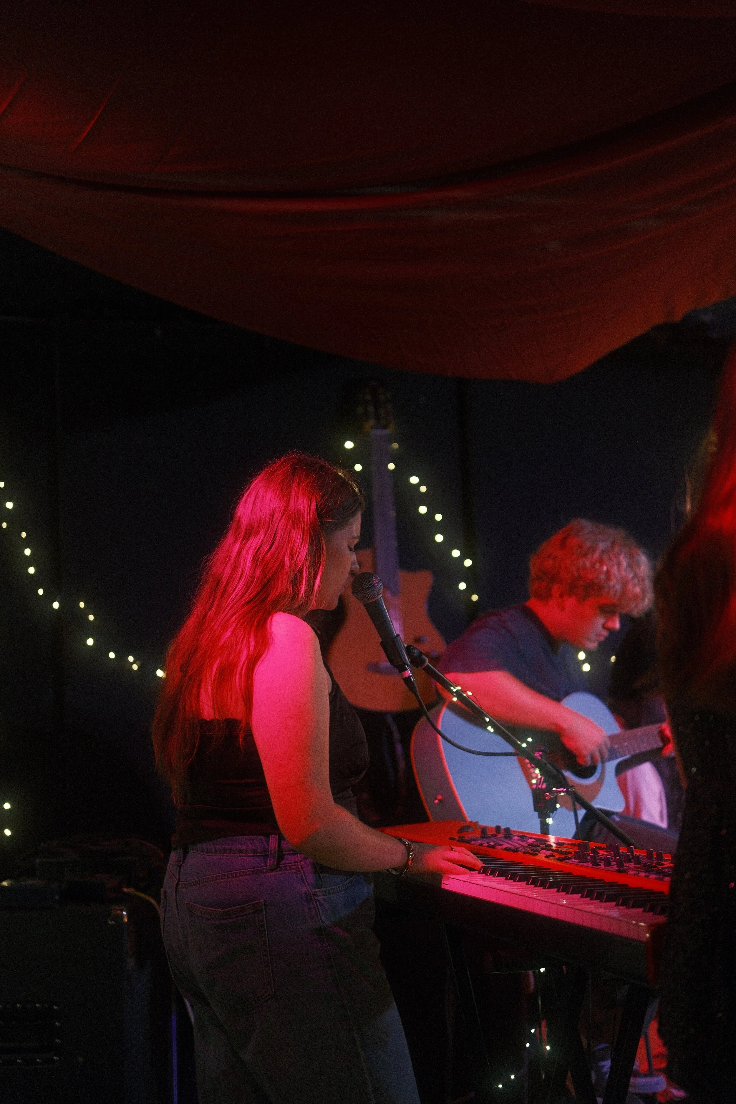 A woman with long red hair playing a keyboard and singing into a microphone on stage, with a man playing an acoustic guitar beside her, under string lights at a live music venue at night.