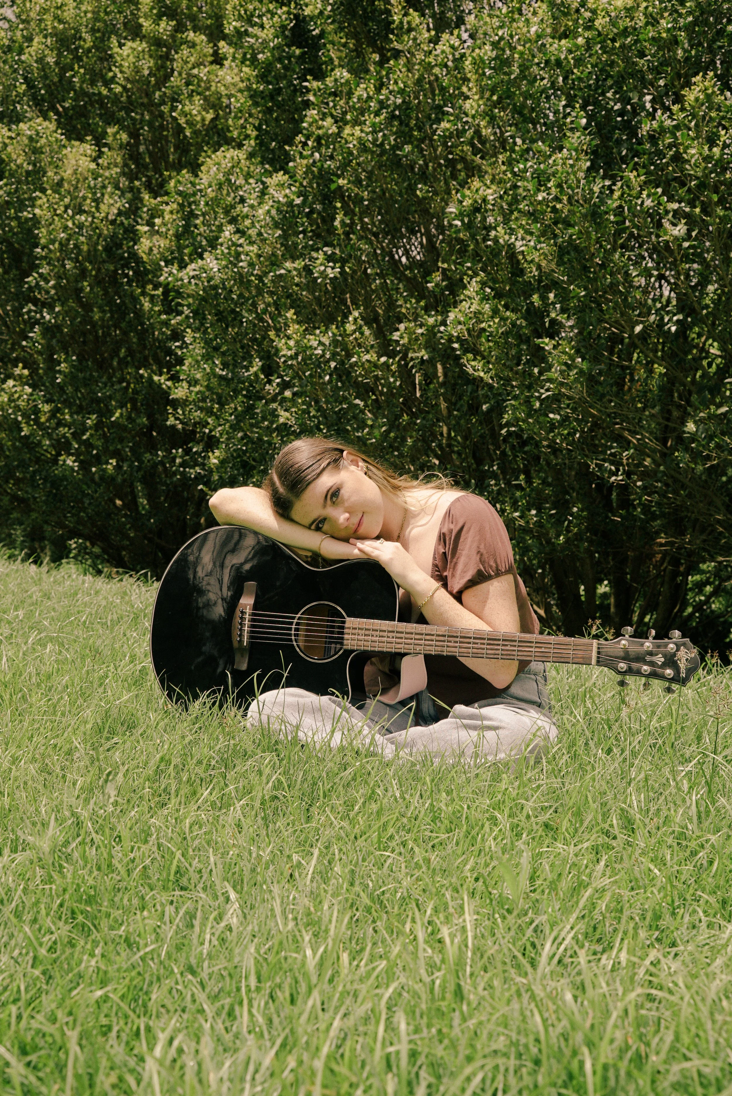 A young woman sitting on the grass playing an acoustic guitar outdoors, with green bushes and trees in the background.
