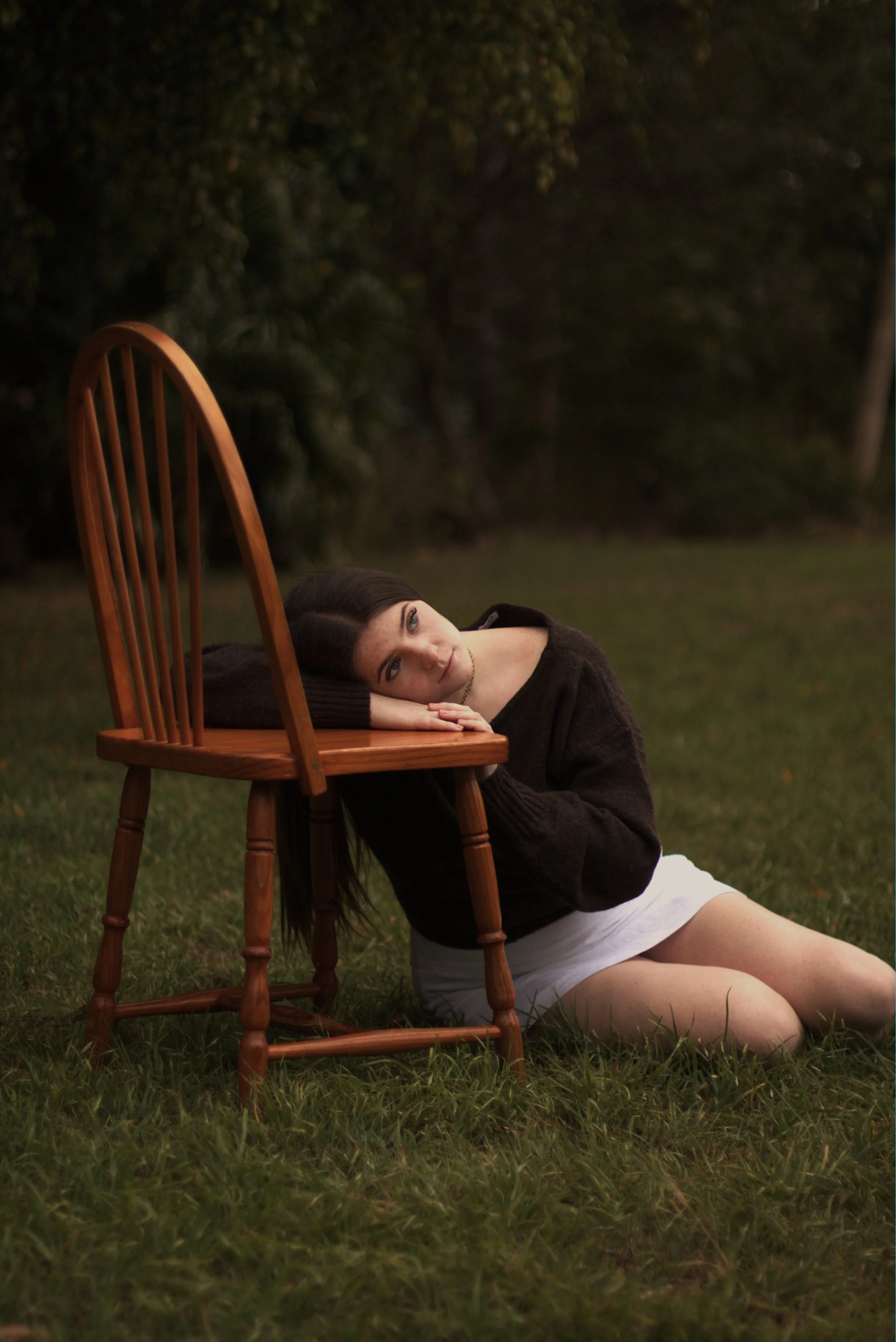 A woman with long dark hair, lying on the grass outdoors, resting her head on her arms on a wooden chair, with a dark, lush background.