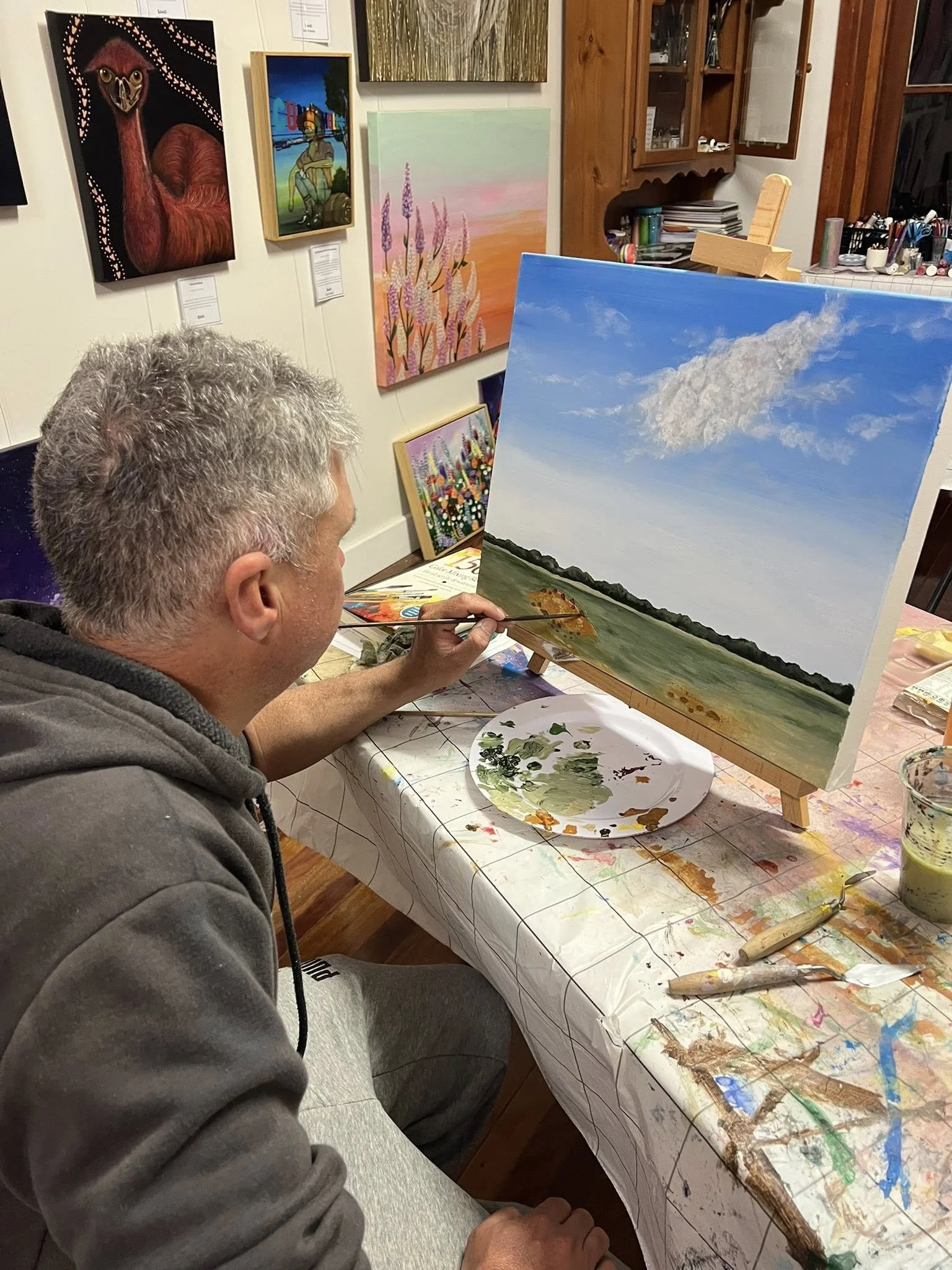A man with gray hair painting a landscape scene of a sky, clouds, and a body of water on a canvas in an art studio, with various paintings and art supplies visible in the background.