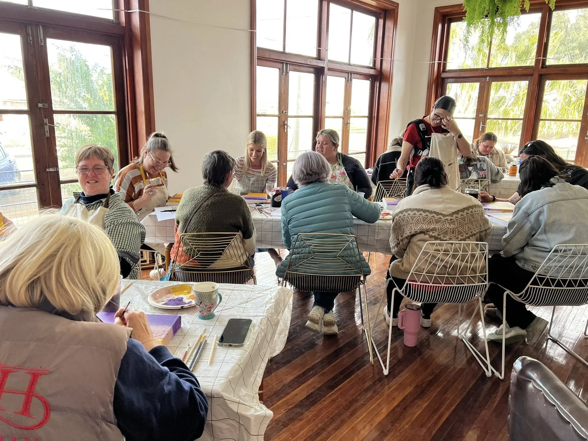 Group of women seated around tables in a well-lit room with large windows, engaged in arts and crafts activities, with some smiling and working on painting projects.