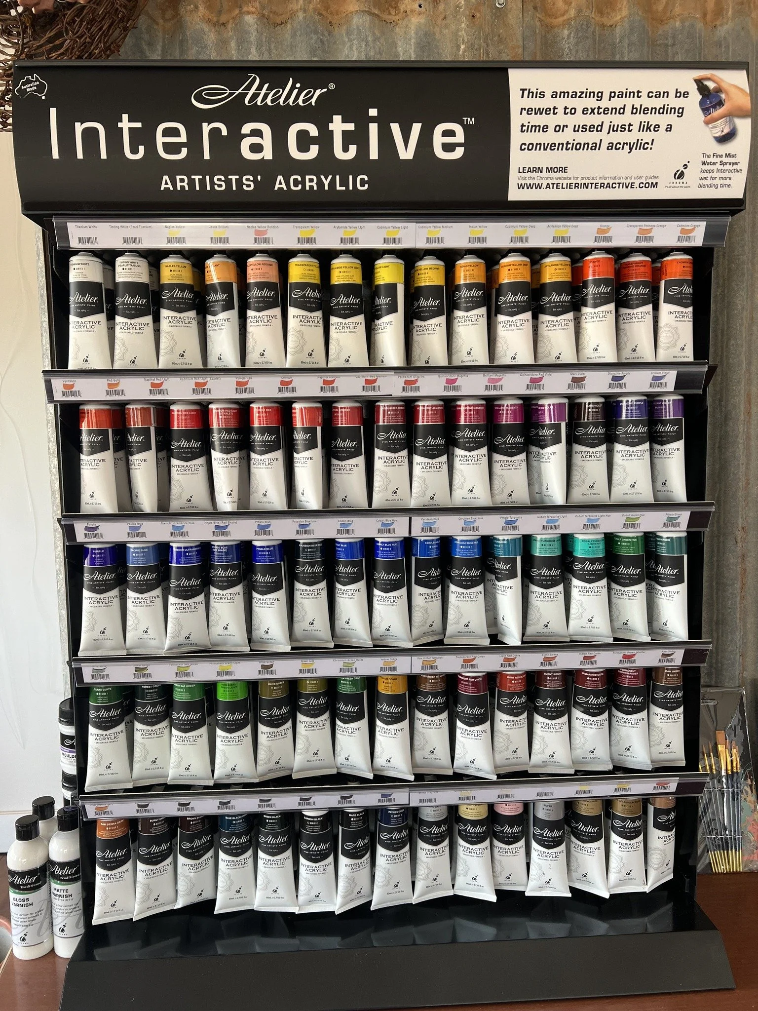 Display of Atelier Interactive artist acrylic paint tubes in various colors arranged on shelves, with bottles of varnish on the side.