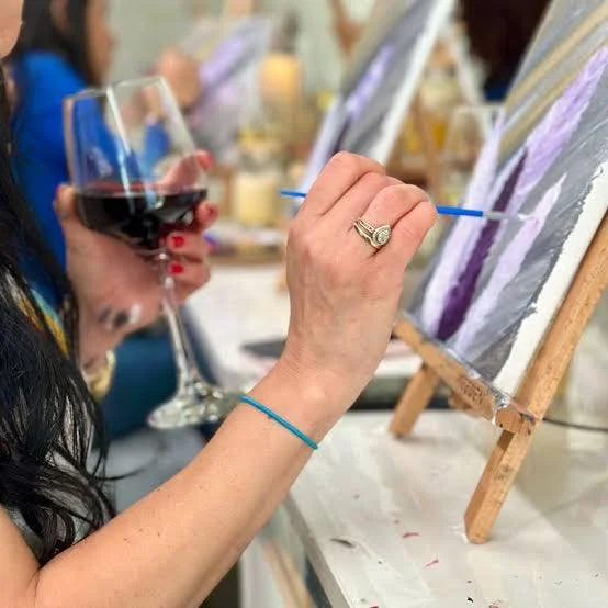 Person painting on canvas at a wine and paint event, holding a glass of red wine.