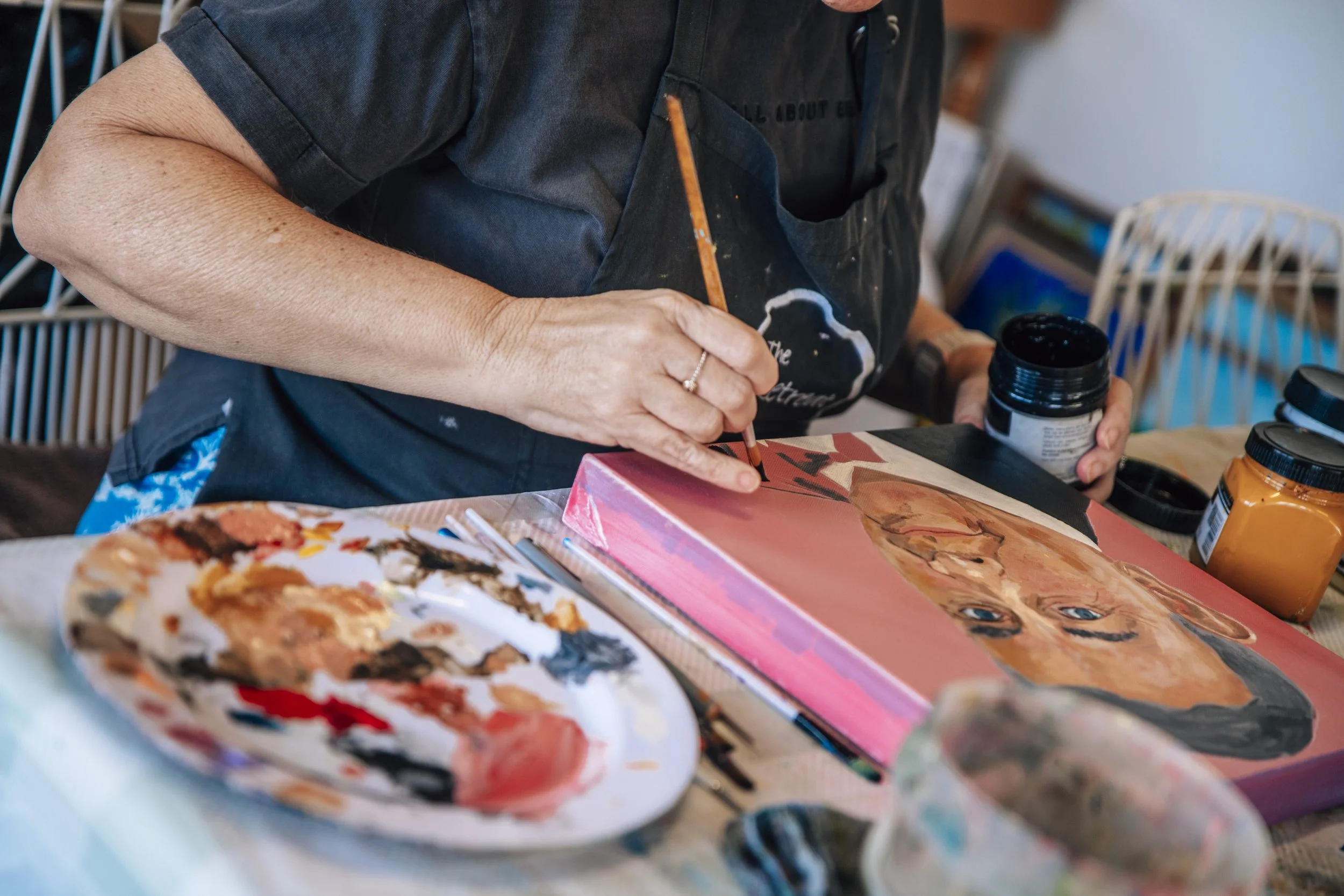 Person painting a portrait on a canvas with various paint jars and brushes on the table.
