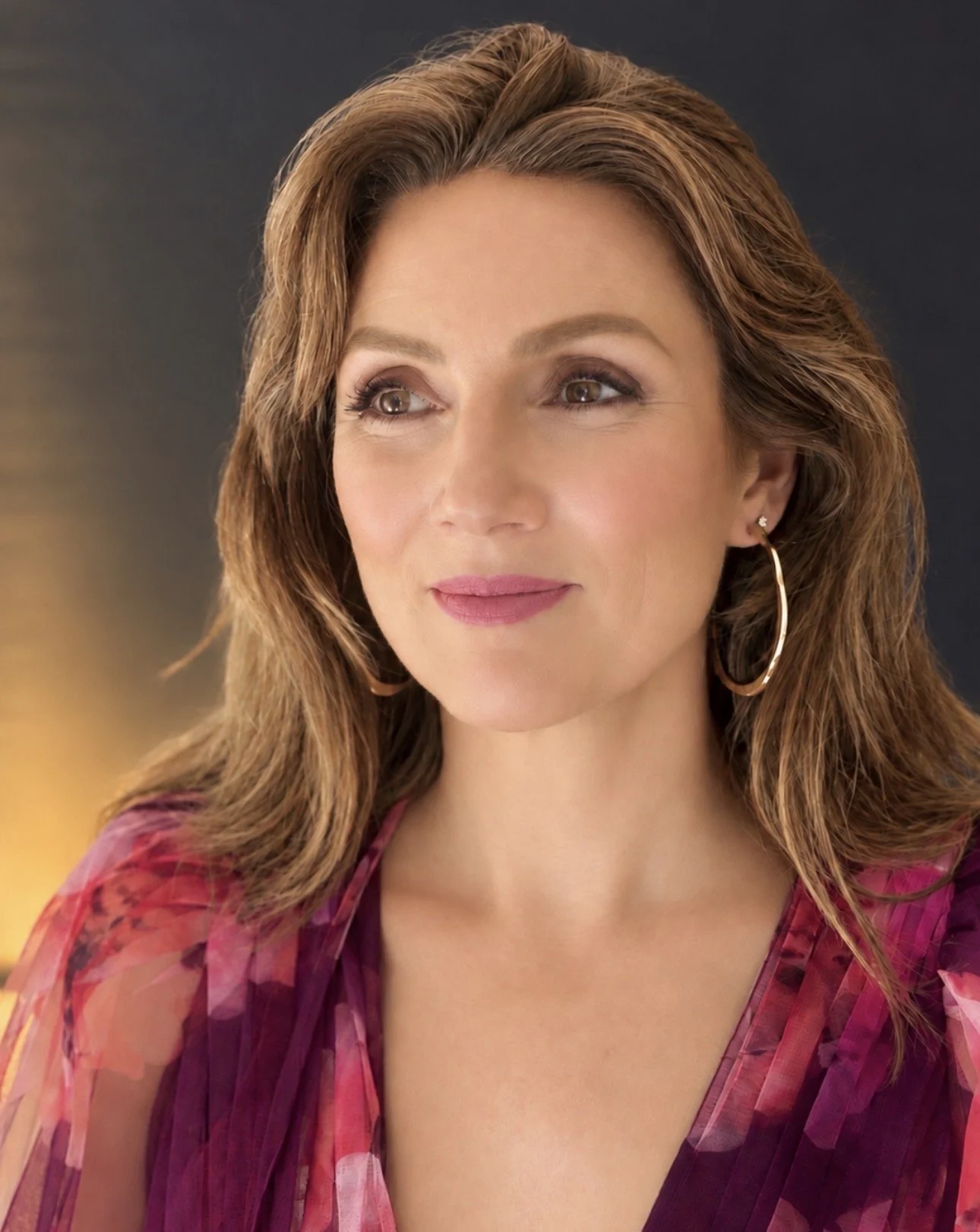 Close-up portrait of a smiling woman with wavy brown hair, hoop earrings, and a pink patterned top