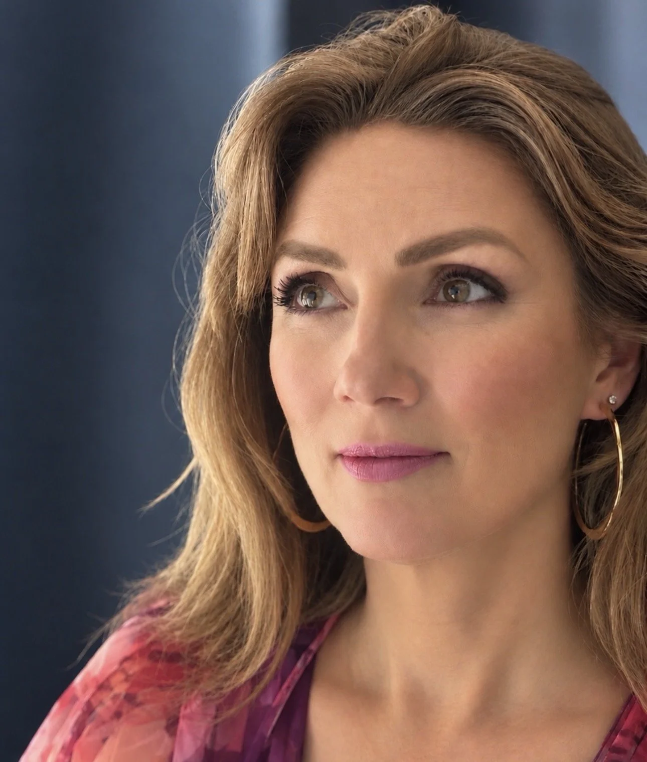 Close-up portrait of a woman with wavy brown hair, green eyes, hoop earrings, and pink lipstick.