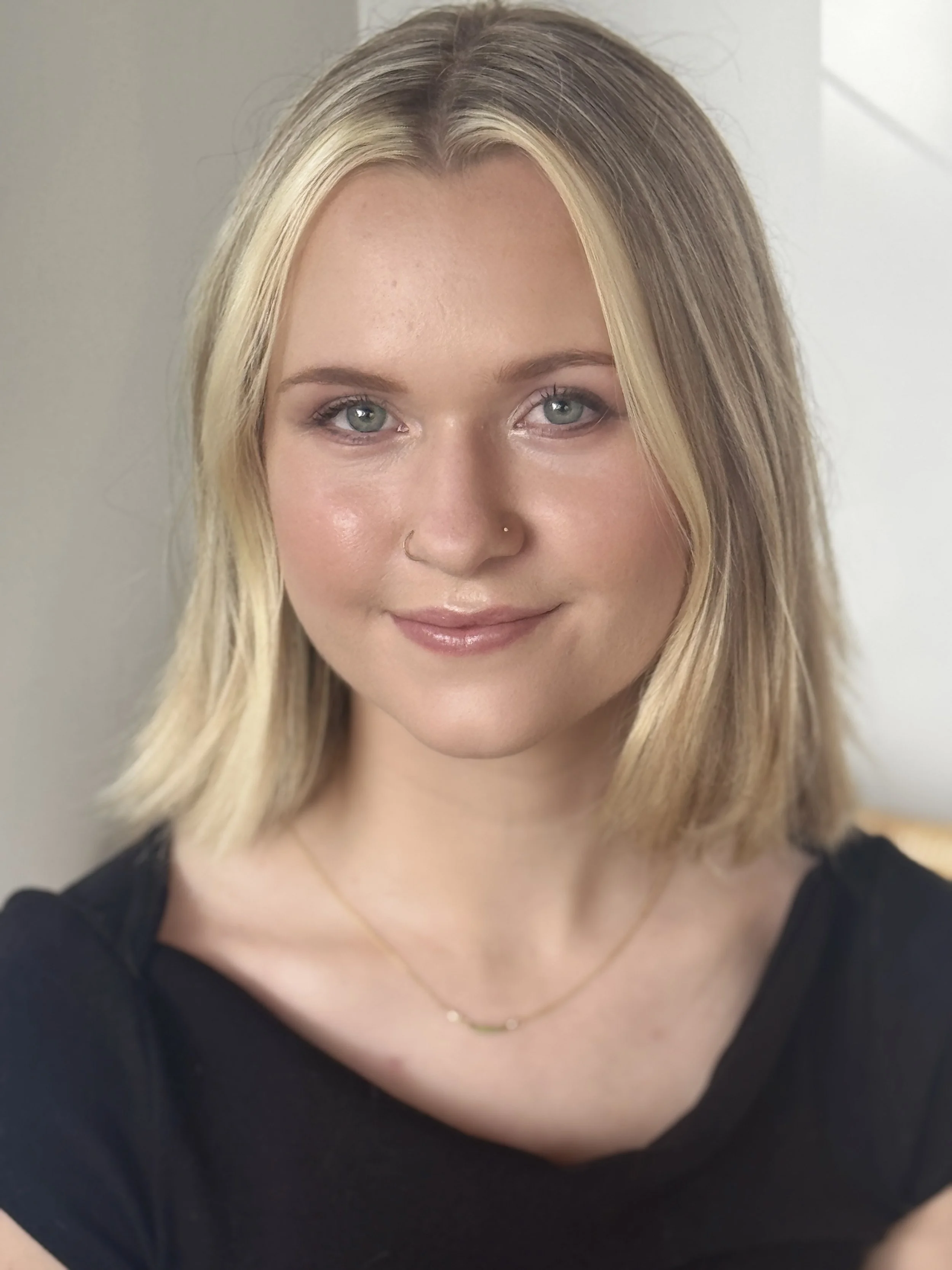 Close-up portrait of a smiling blonde woman with green eyes, a nose ring, and a delicate necklace.