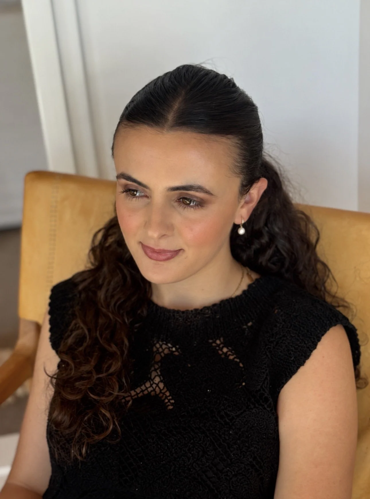 Portrait of a woman with dark curly hair wearing a black knit top and pearl drop earrings, seated in a mustard-yellow chair.