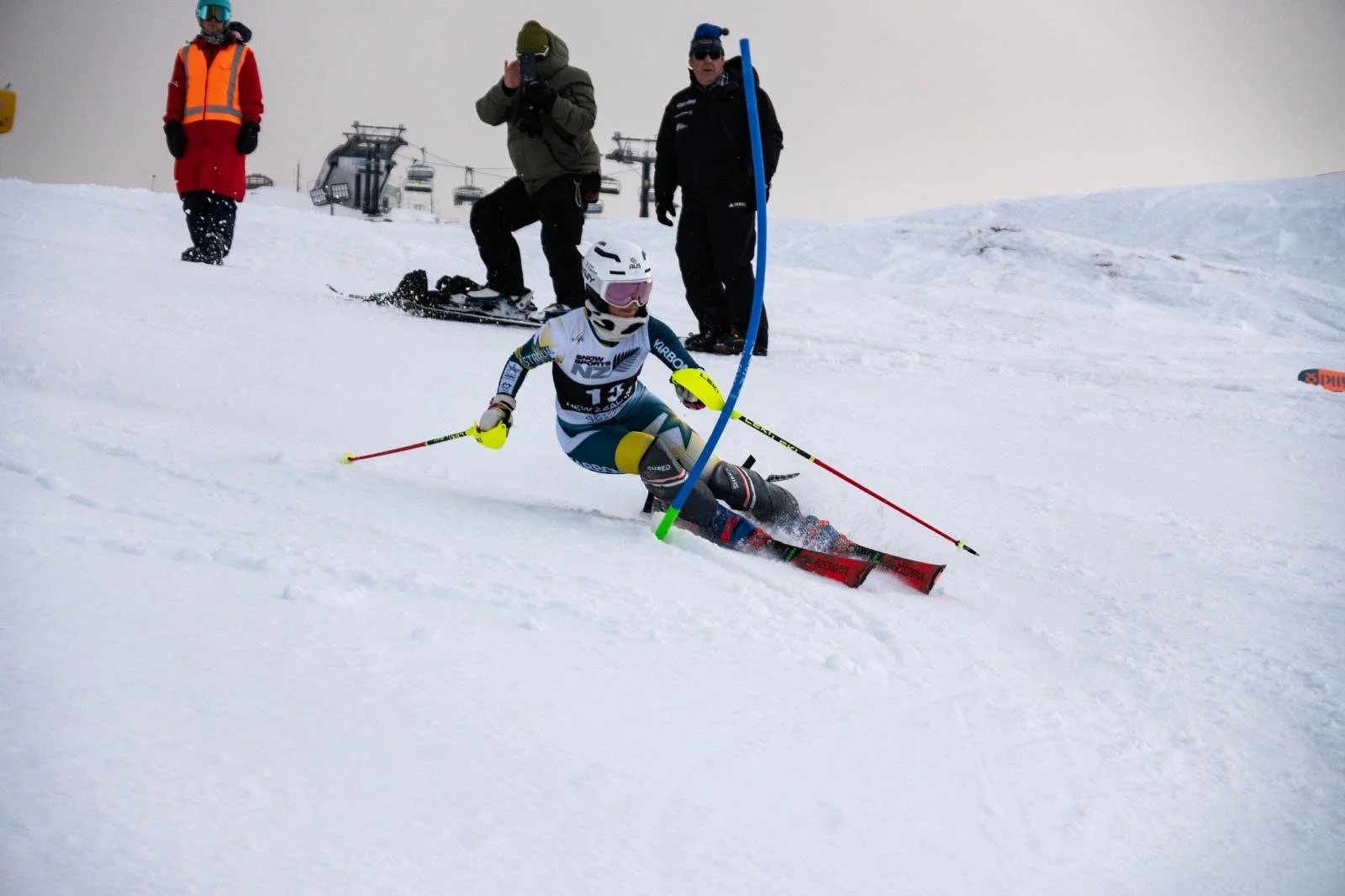 Competitive skier Madi Gilson, in the Australian national team race suit, speeding around a blue slalom gate on a snowy slope, with spectators and ski lift chairs in the background.