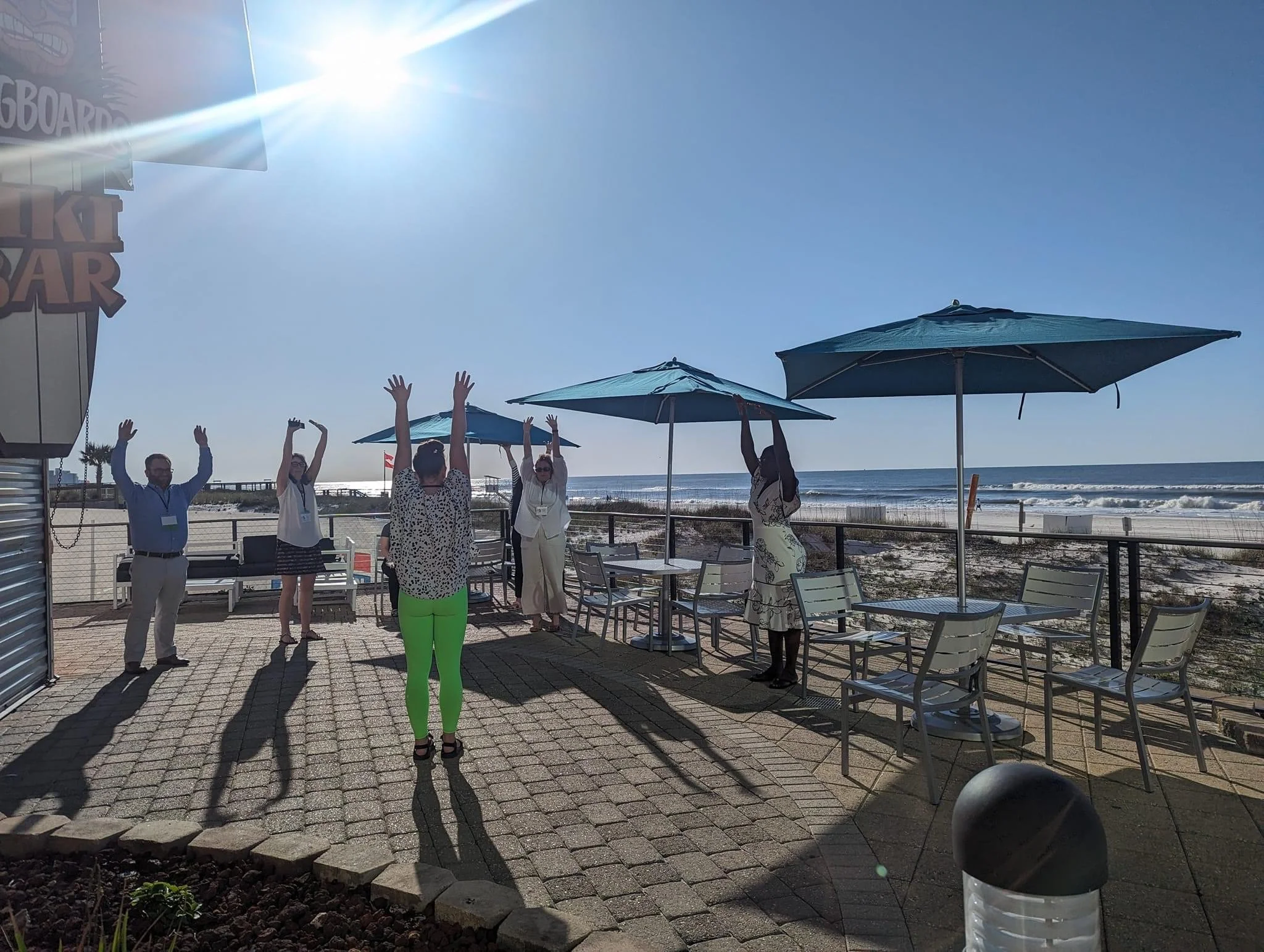 Group of people raising their hands on a beachfront patio beneath umbrellas, with ocean waves and a sunny blue sky behind them