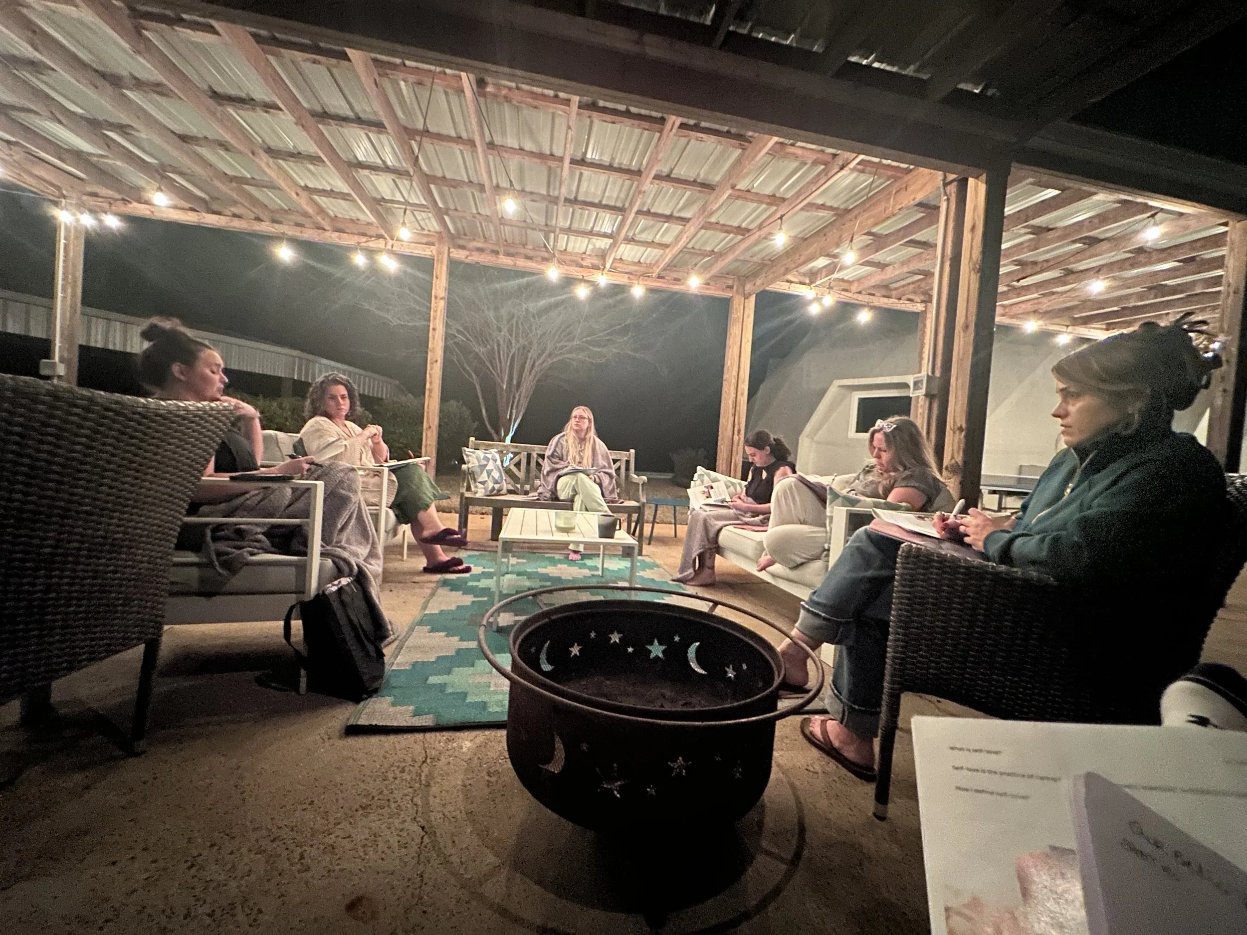 Group of people sitting in a covered outdoor patio at night, writing in notebooks around a fire pit under string lights.