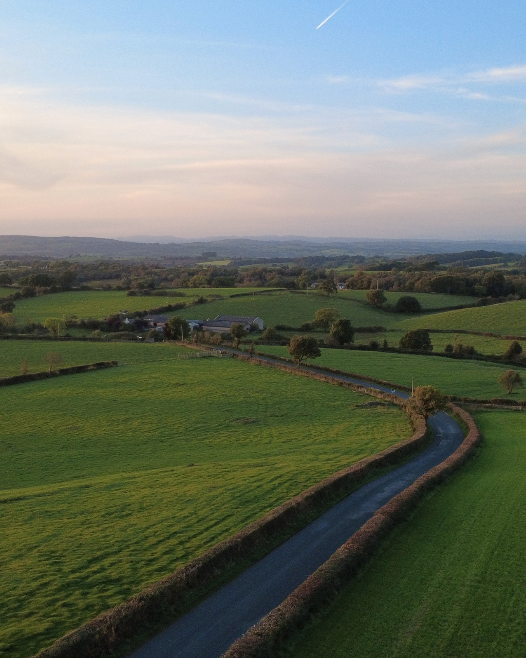 Aerial view of rolling green countryside with fields separated by hedgerows and a winding narrow road, under a sky with some clouds and a jet streak.