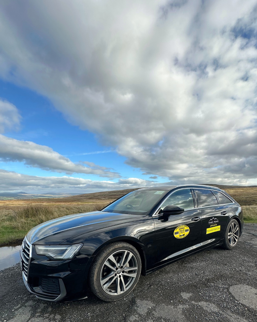 Black Audi A6 taxi parked on a gravel road in a rural area with grassy fields and hills under a cloudy sky.