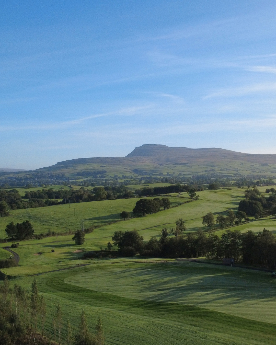 A lush green golf course with rolling hills and trees, with a mountain in the background under a blue sky with wispy clouds.