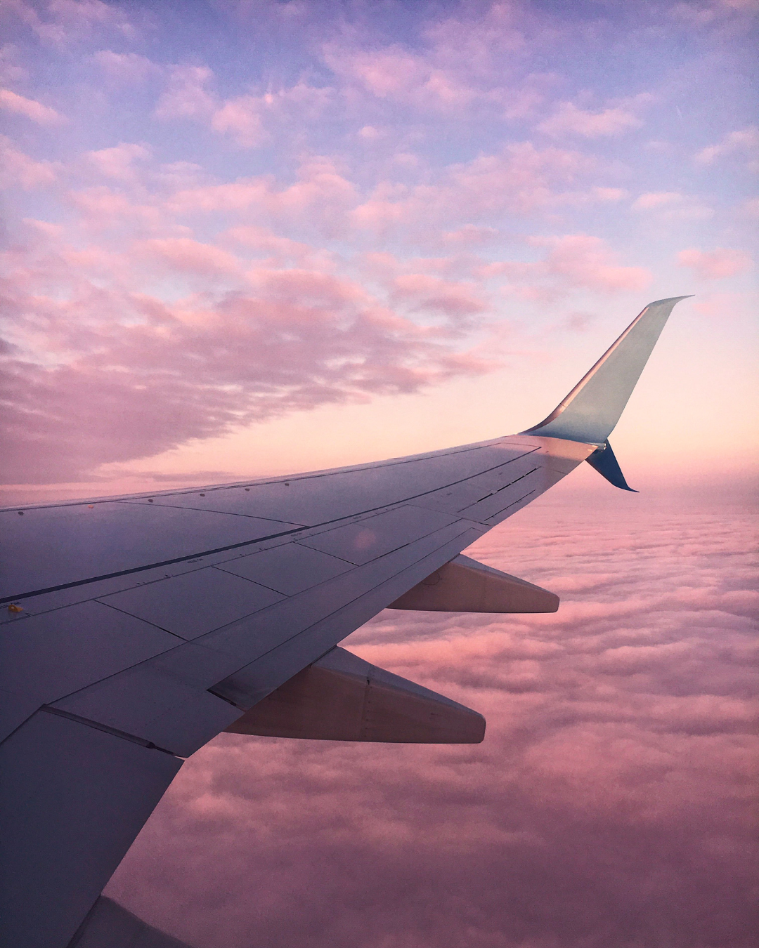 View of airplane wing flying above pink and purple clouds at sunset or sunrise.