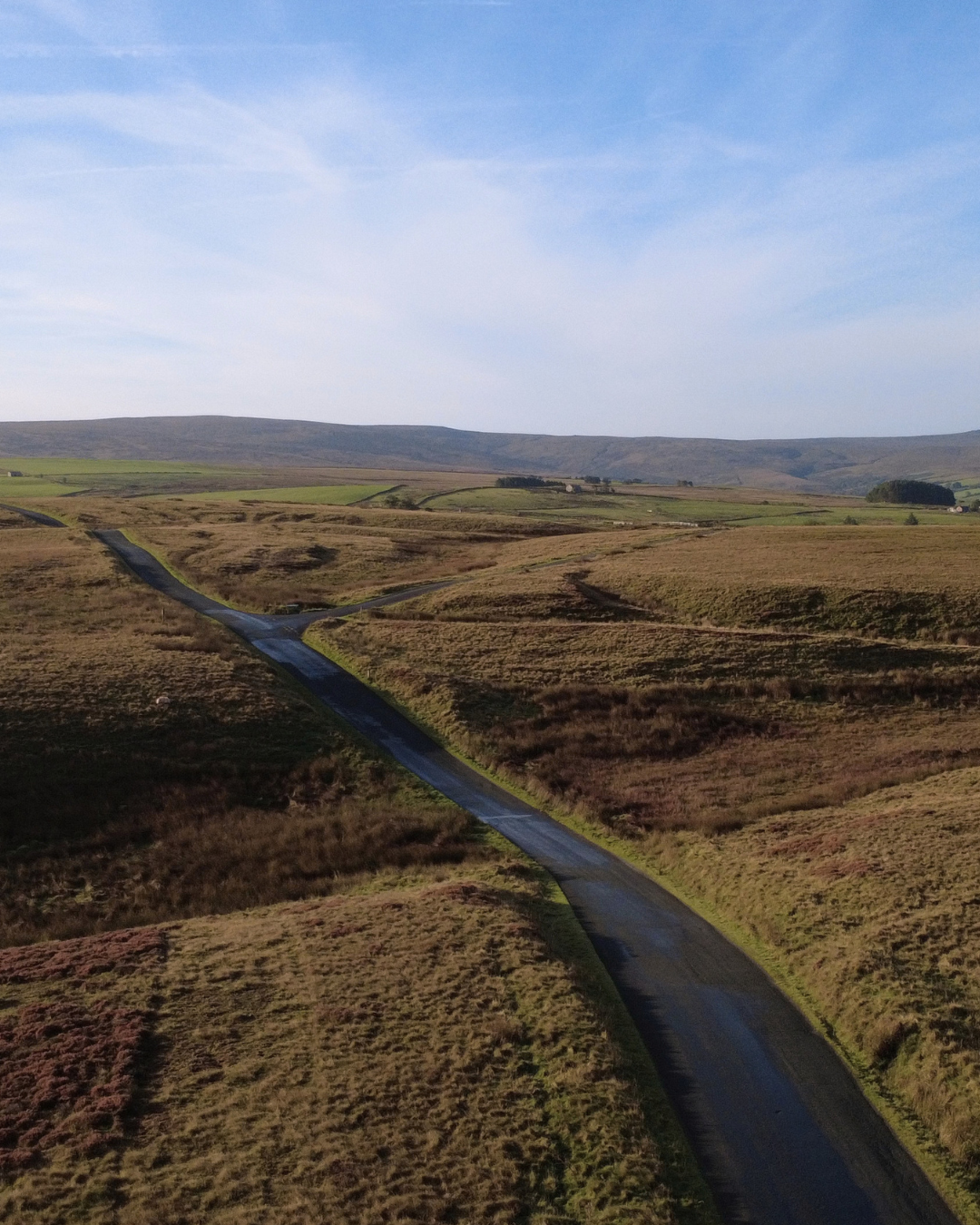 A winding road across open grassy fields with hills in the background under a blue sky with wispy clouds.
