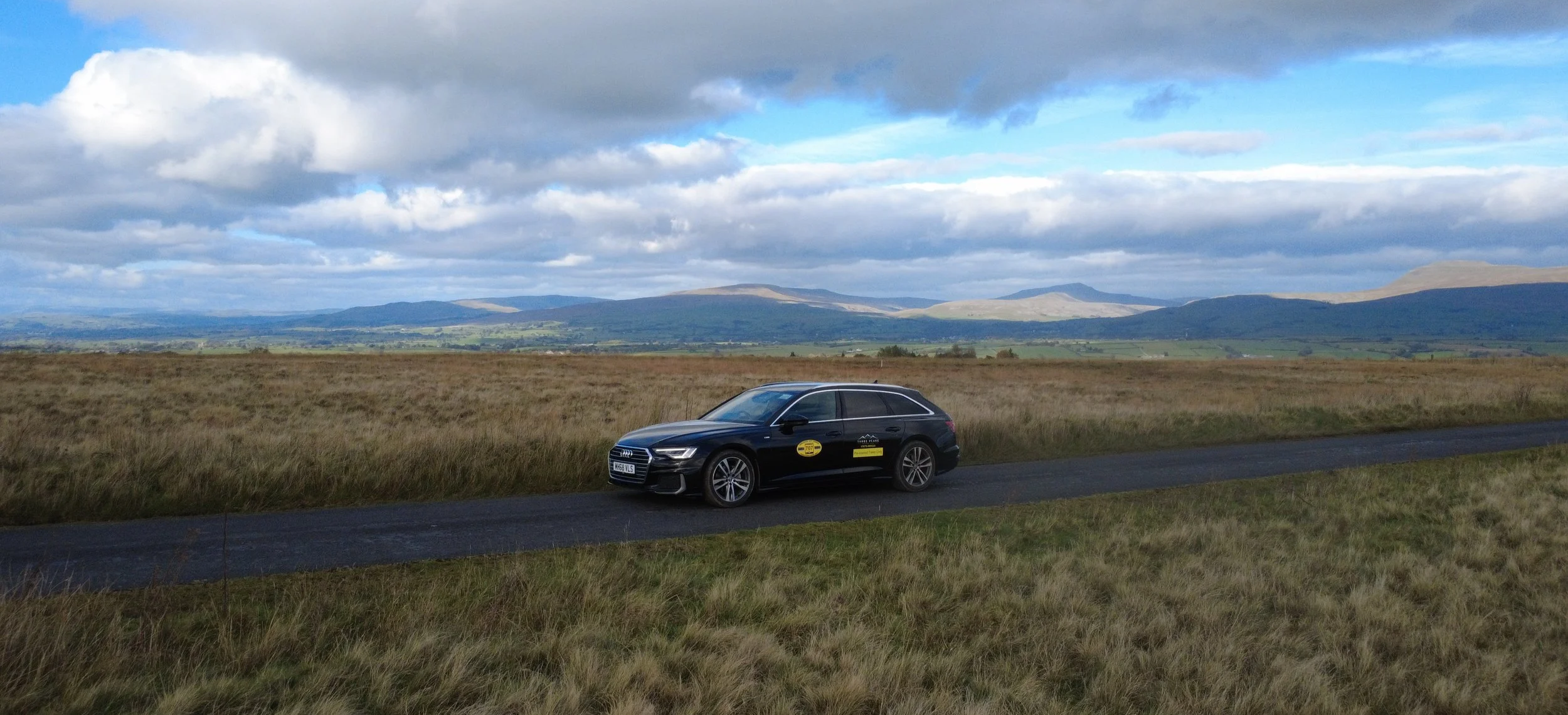 A black Audi car with a yellow sign on the door parked on the side of a rural road in a grassy field, with distant rolling hills and a partly cloudy sky in the background.