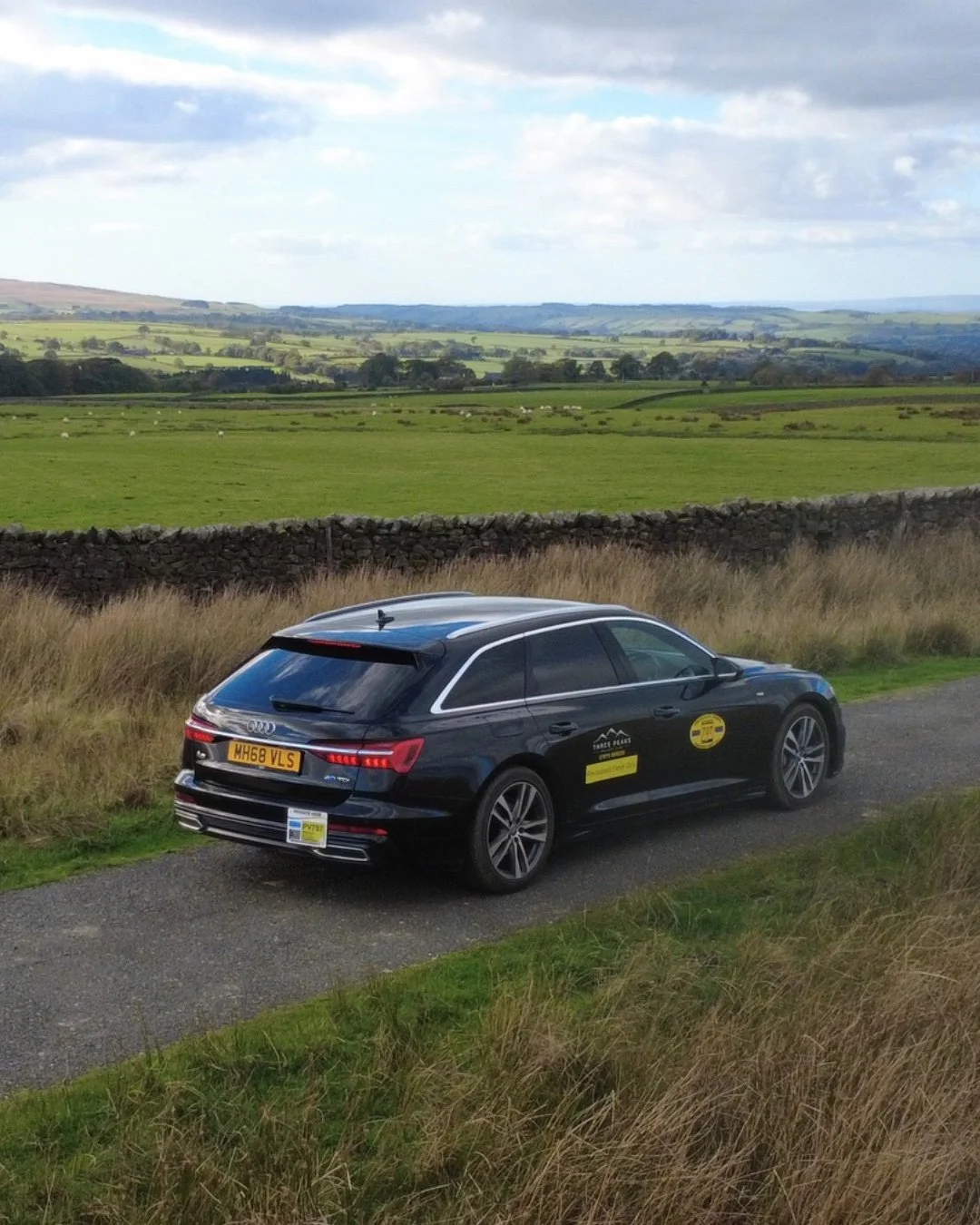 A black Audi A6 taxi from Three Peaks Private parked on a narrow gravel road amidst green fields and rolling hills under a cloudy sky.