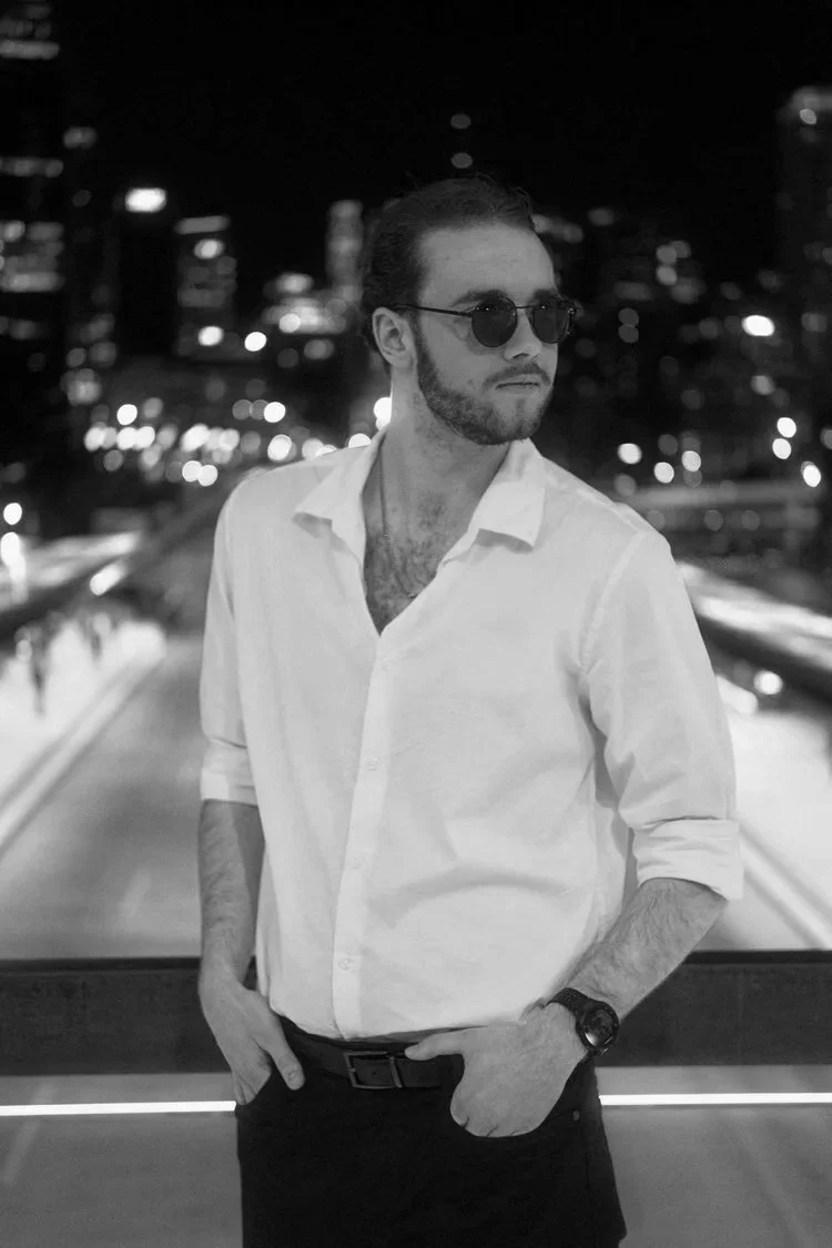 Harry standing on a bridge at night in Brisbane's urban areas, wearing a white shirt with rolled-up sleeves and dark pants, with city lights and buildings blurred in the background.