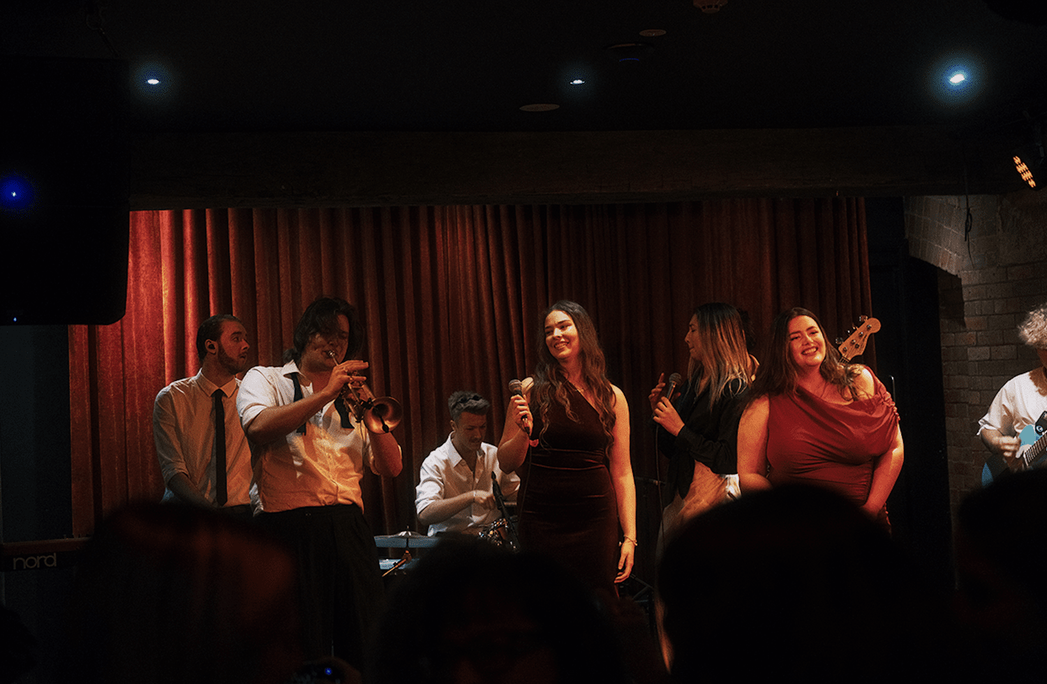 'The Contraband' perform on stage in a dimly lit venue with a red curtain backdrop. The vocalists are holding microphones, others are playing instruments, and they are smiling and engaging with the audience.