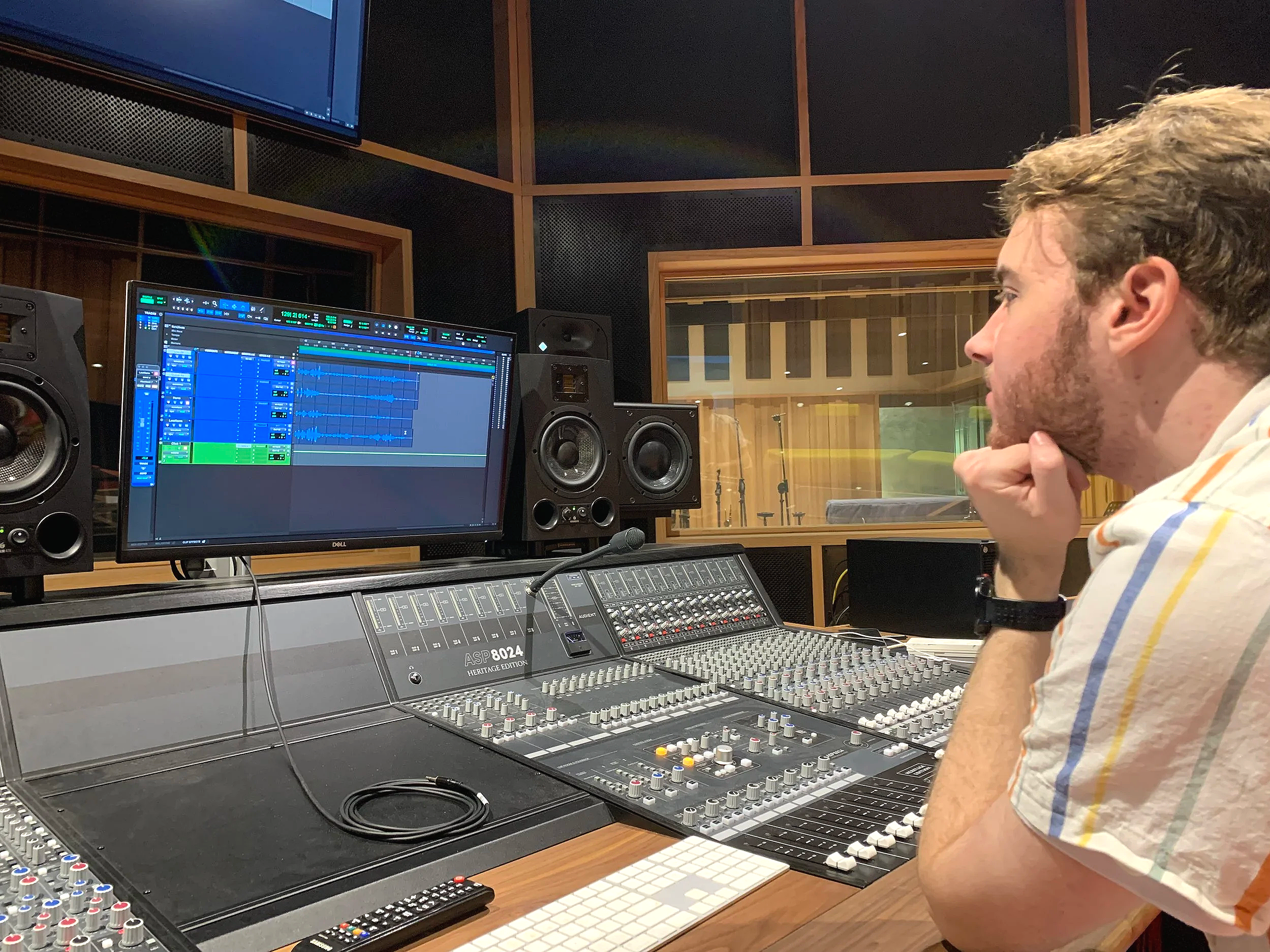 A man with curly hair and a beard sitting in a recording studio, looking at a computer screen displaying a digital audio workstation, with various audio equipment and a mixing console on the desk.