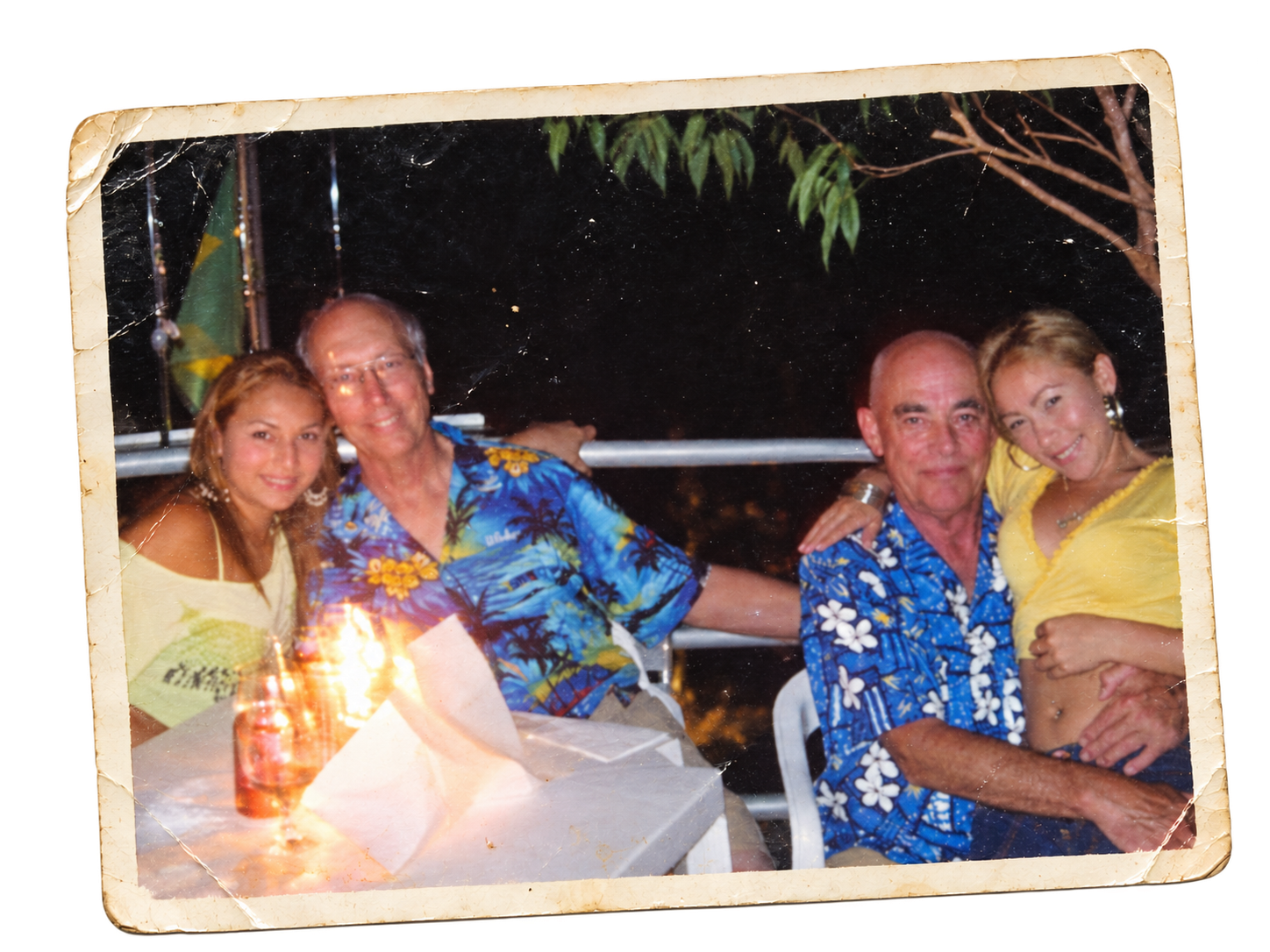 A family of five posing at night, sitting at a table outdoors. The group includes a young woman, two middle-aged men, and two middle-aged women, all wearing brightly colored Hawaiian shirts or tops. There is a bottle on the table, and the background shows dark night sky with some trees and a flag.