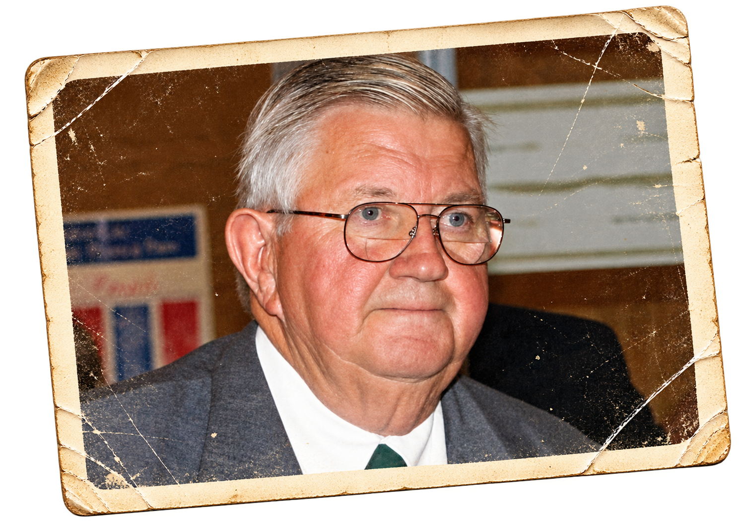 A close-up photograph of an older man with gray hair, wearing glasses, a white shirt, a green tie, and a dark jacket, sitting in front of a wood-paneled background with some posters. The photo appears old and weathered with visible creases and tears, and is framed with a worn, torn corner.