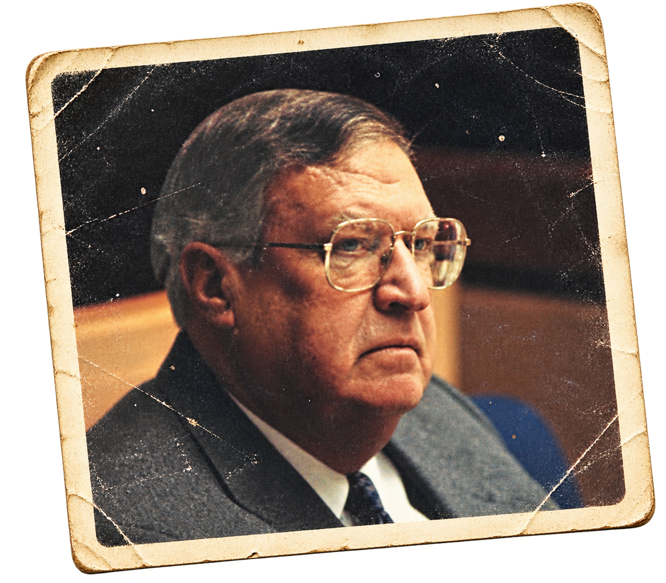 An old, worn photograph of a stern-looking man with glasses, wearing a suit and tie, with a wooden background and a black backdrop.
